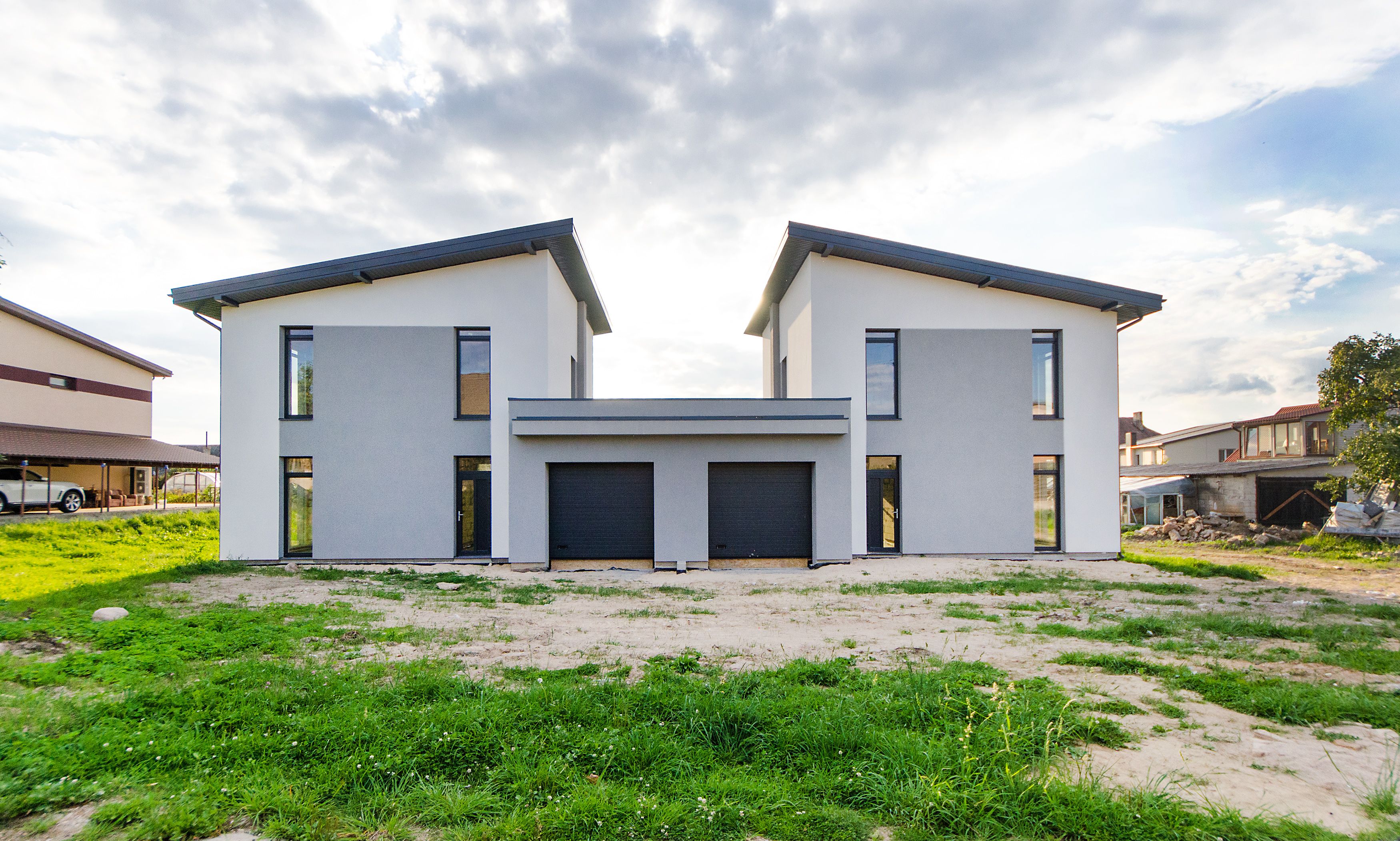 A modern twin-house with white and grey stucco, black trim, and two charcoal garage doors, set on a manicured lot under a clear sky.