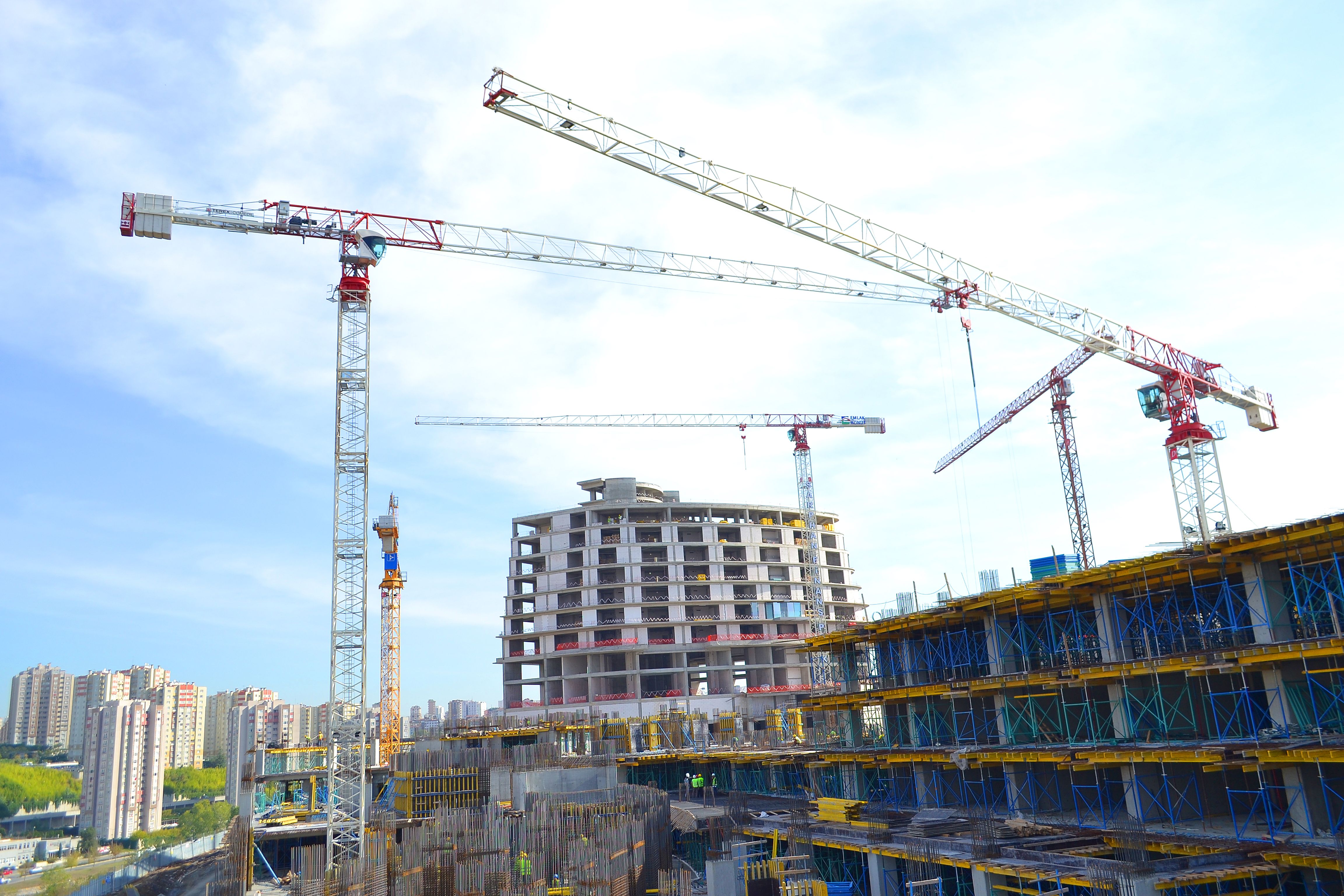 Three red-and-white tower cranes rise over a curved concrete high-rise under construction against a clear sky.
