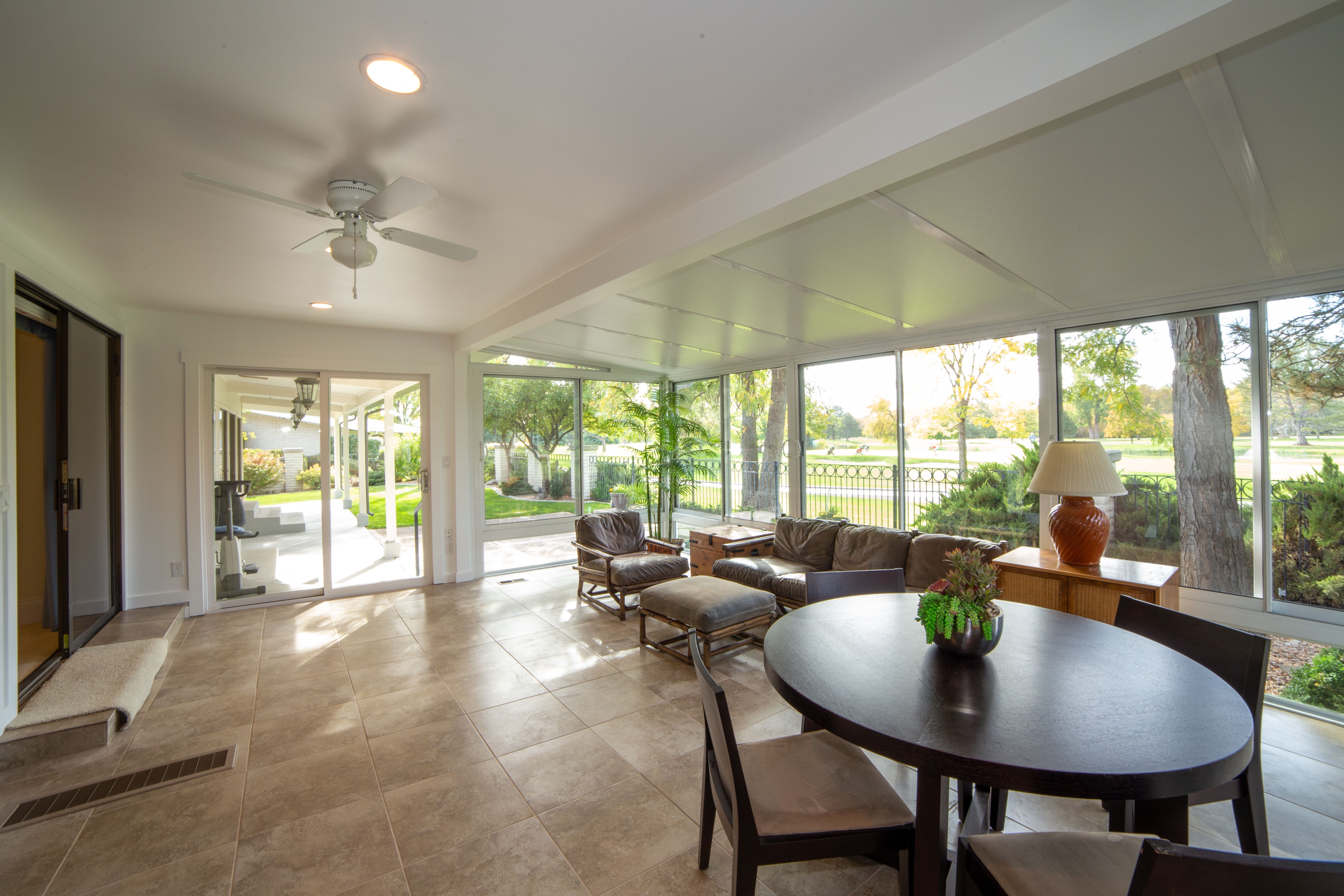 A finished home sunroom addition with floor-to-ceiling glass windows, a leather sofa set, a round dining table, and a sliding door opening onto a covered patio with a view of trees and a stone-arch bridge.