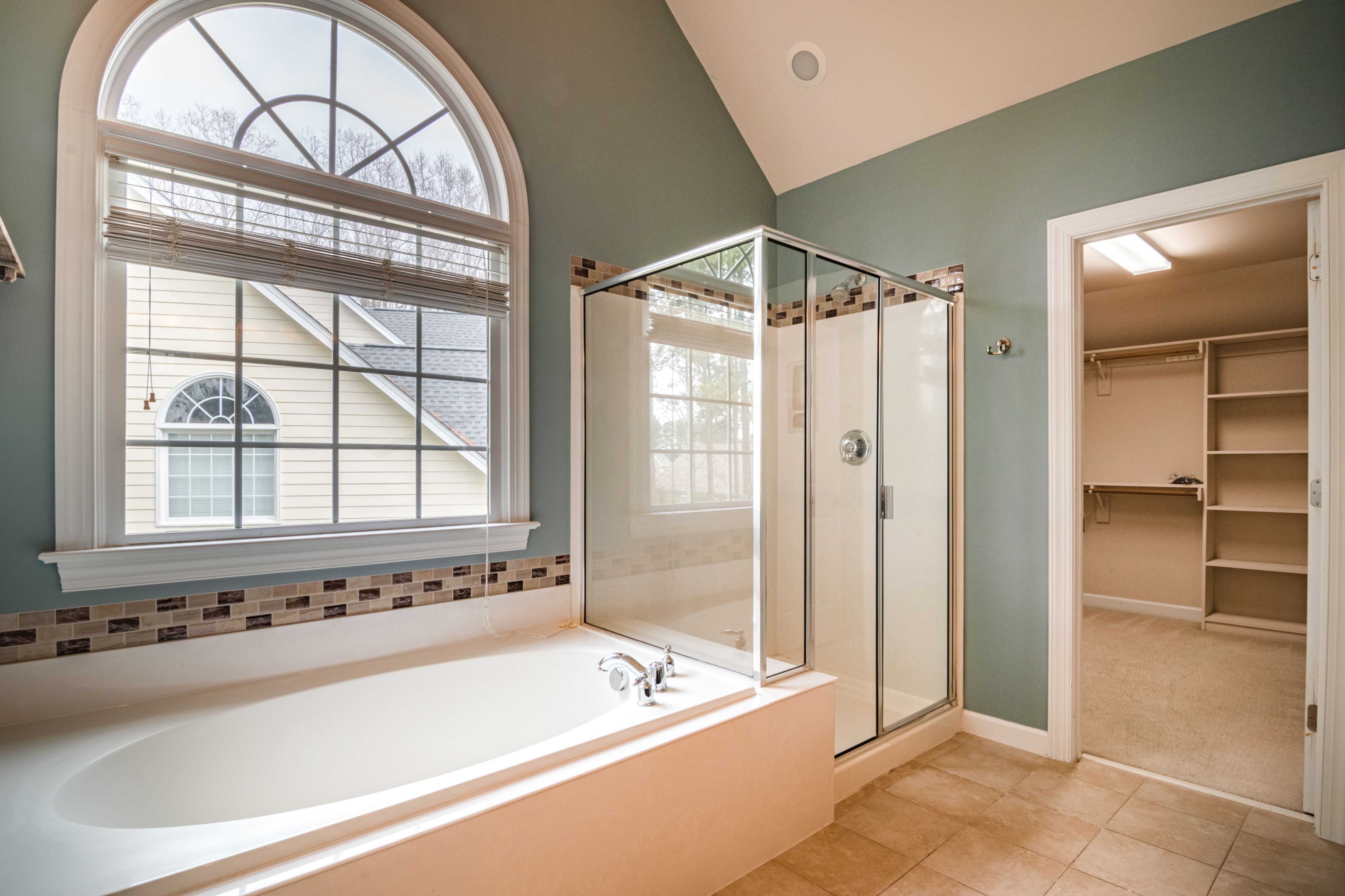 A finished luxury master bathroom with a deep soaking tub under an arched Palladian window, a glass-enclosed shower, soft sage-green walls, and an open doorway to a walk-in closet.