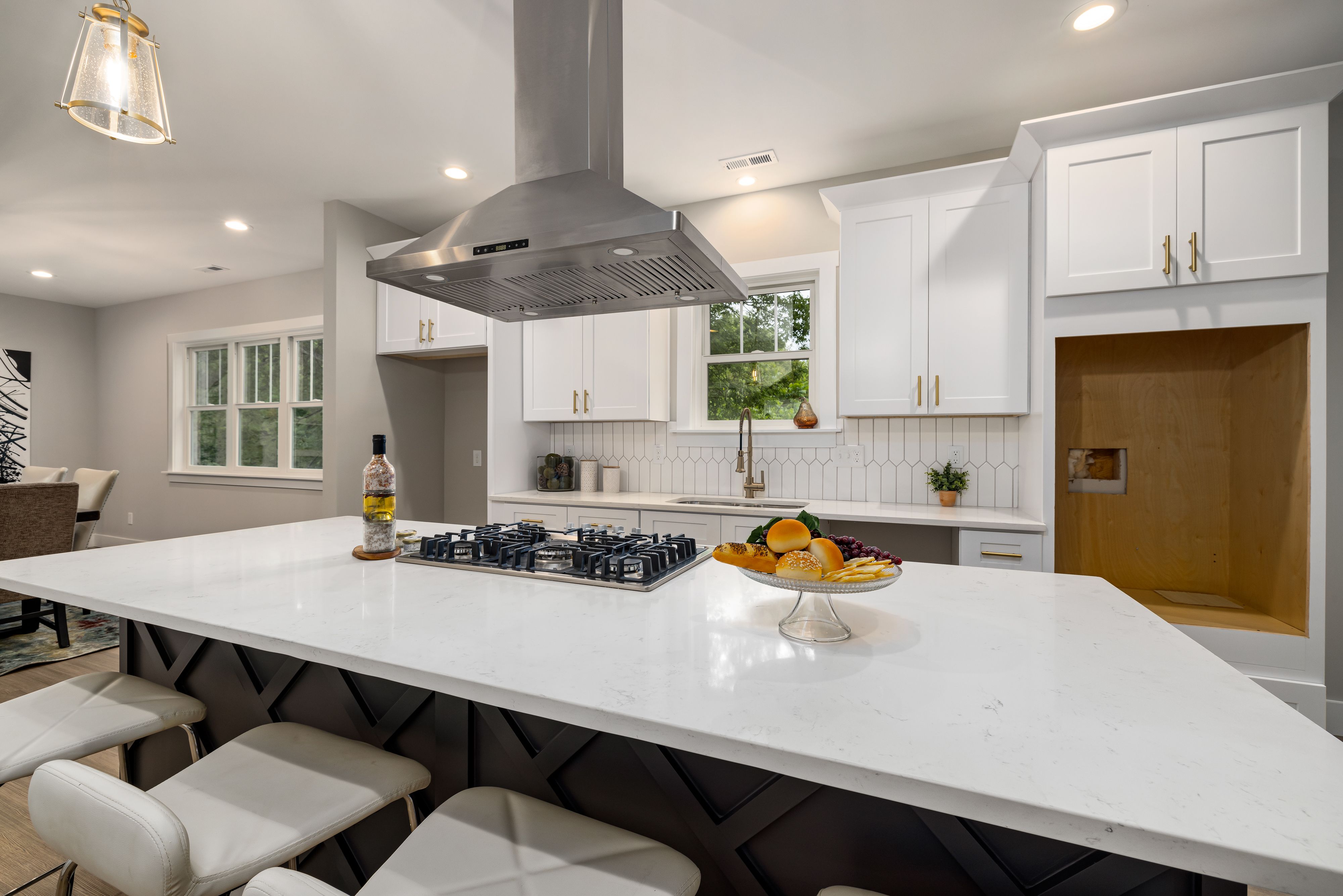 A finished modern kitchen with a large white quartz waterfall island, black cooktop, stainless range hood, and white Shaker cabinets with gold hardware.