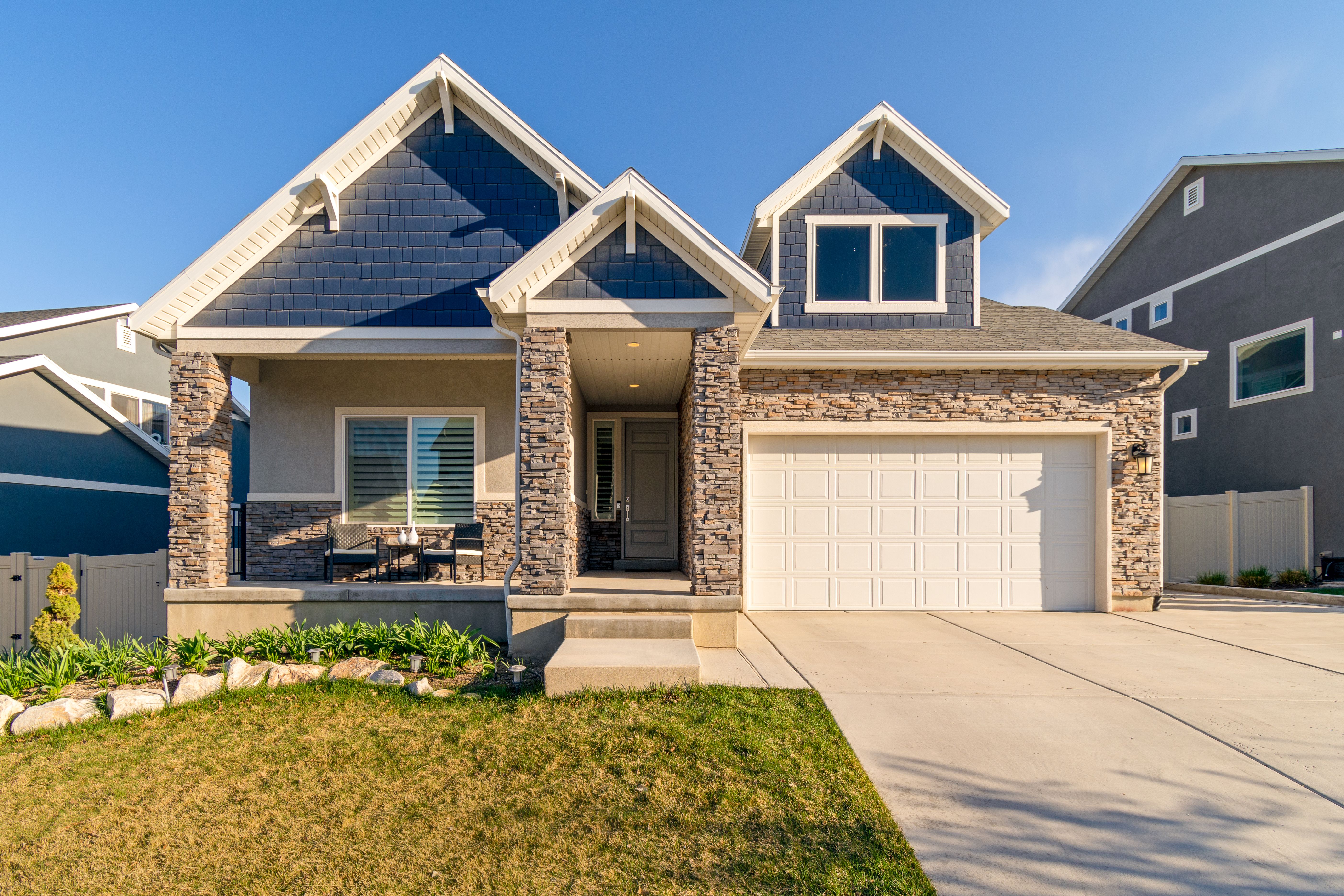 A finished two-story craftsman-style suburban family home with navy-blue shingle siding, a stacked-stone facade, a covered front porch, and a two-car garage, set on a green lawn under a clear blue sky.