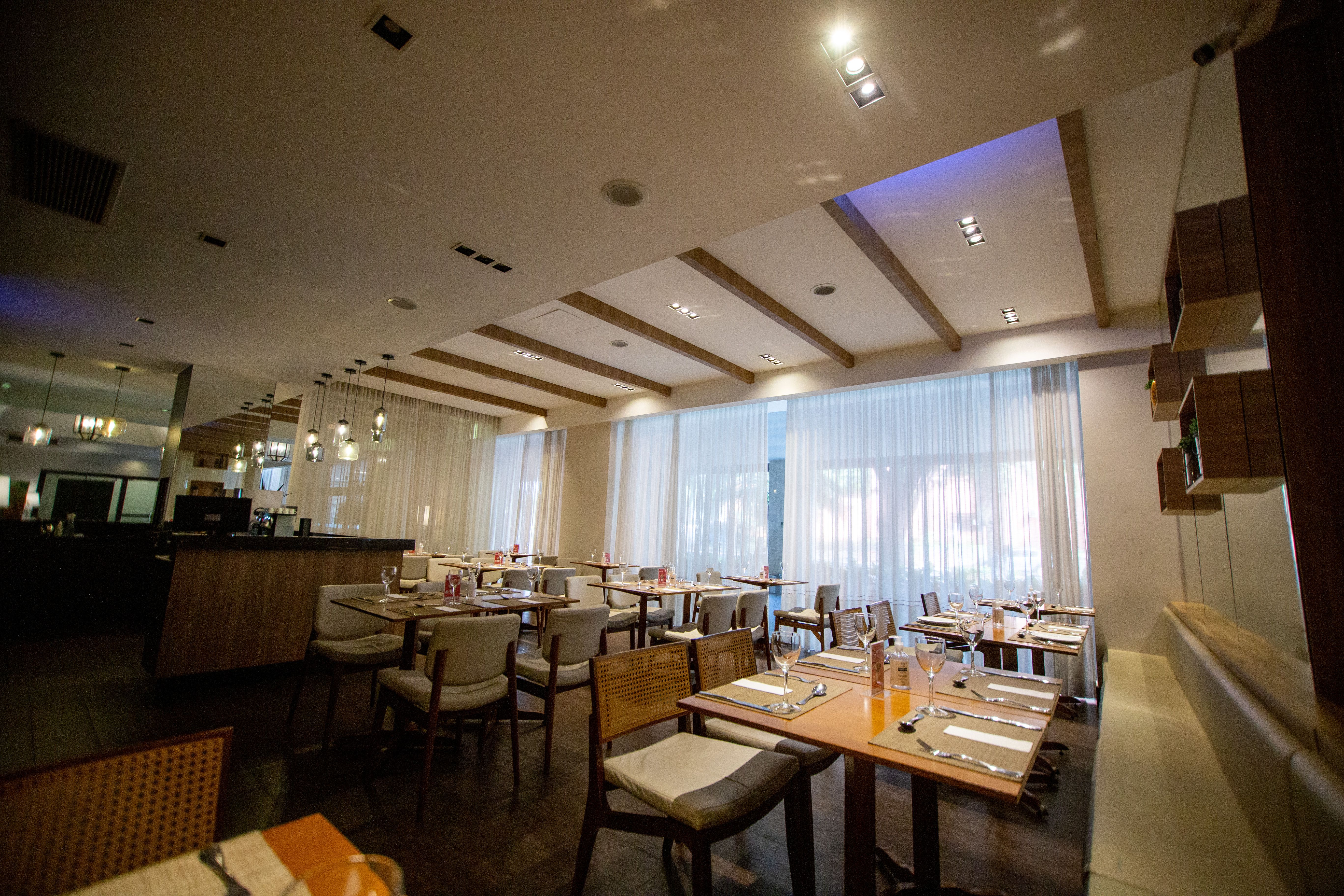 A finished modern restaurant dining room with warm pendant lights, dark-wood tables set with linen placemats, cream upholstered chairs, and sheer floor-to-ceiling curtains along the back window wall.