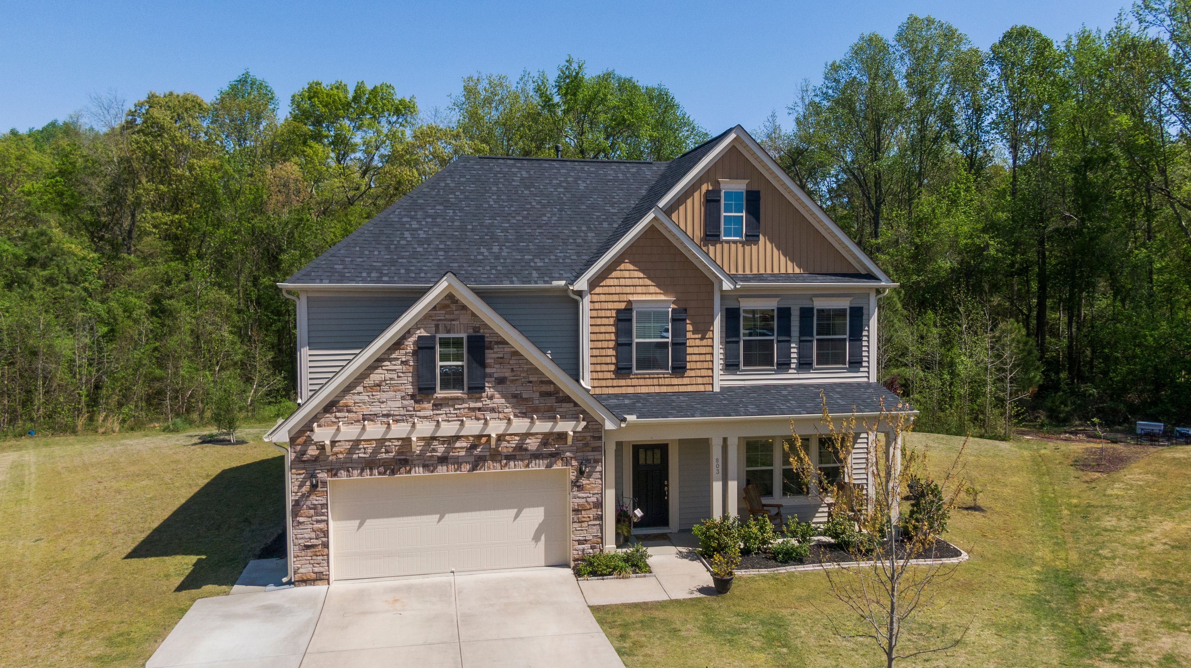 A finished two-story suburban home with a clean charcoal-gray architectural-shingle roof, stacked-stone facade over the garage, board-and-batten cedar gable, and a manicured green lawn under a clear blue sky.