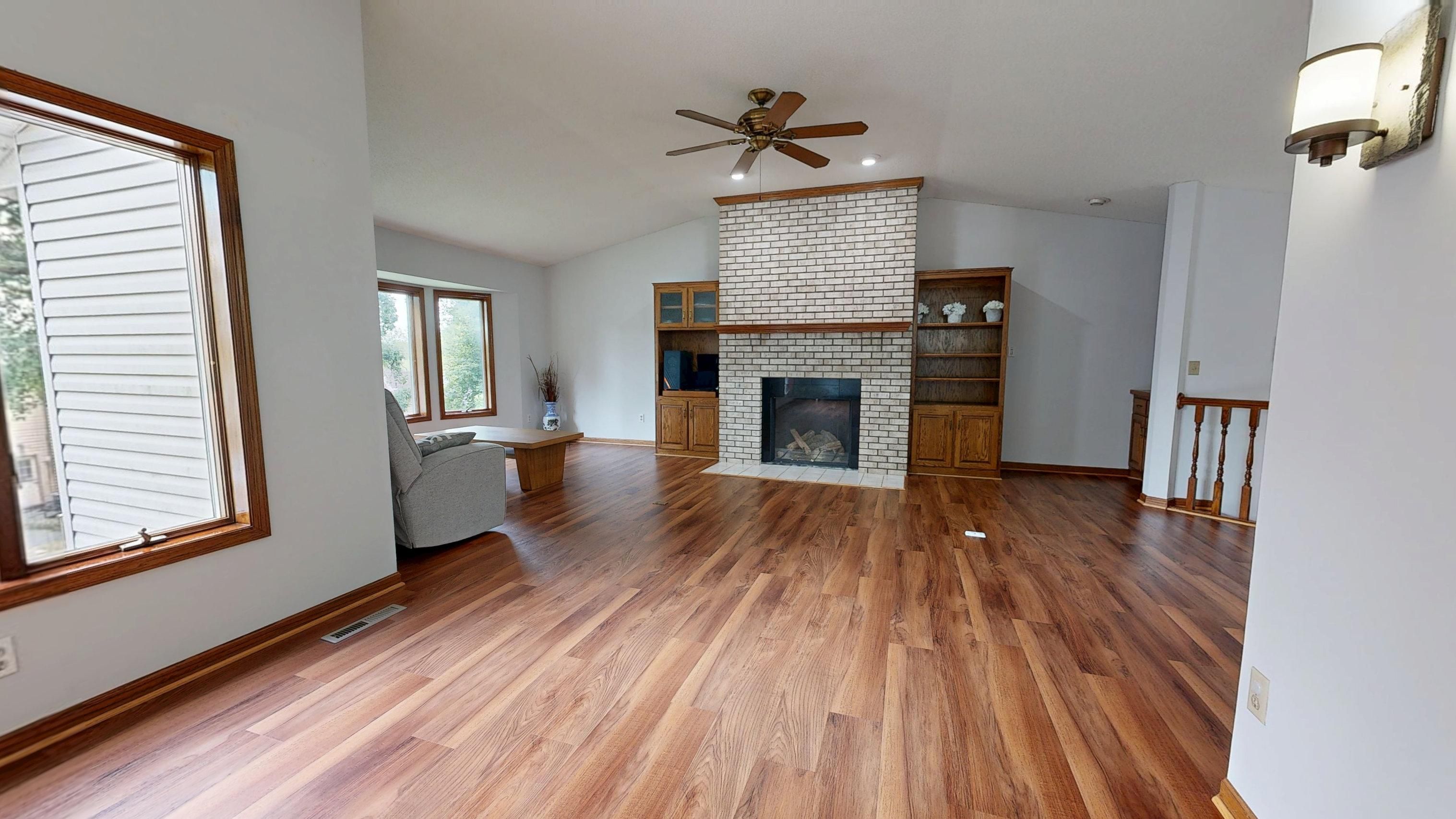 A finished remodeled living room with newly refinished hardwood floors, a floor-to-ceiling white-painted brick fireplace, a vaulted ceiling with a ceiling fan, and built-in oak shelving.