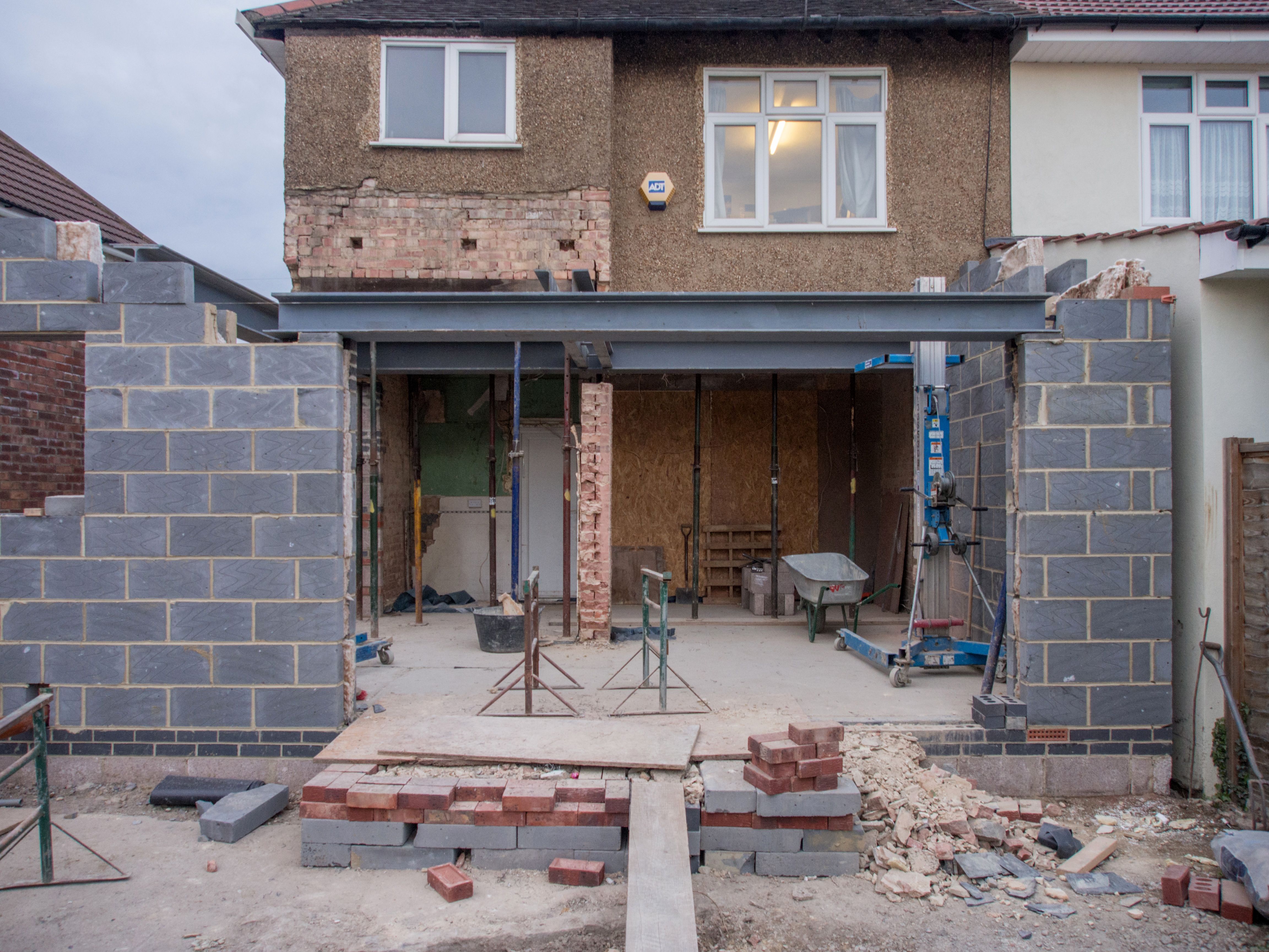 A semi-detached brick house undergoing a rear addition with new concrete block walls, steel beams, and shoring props.