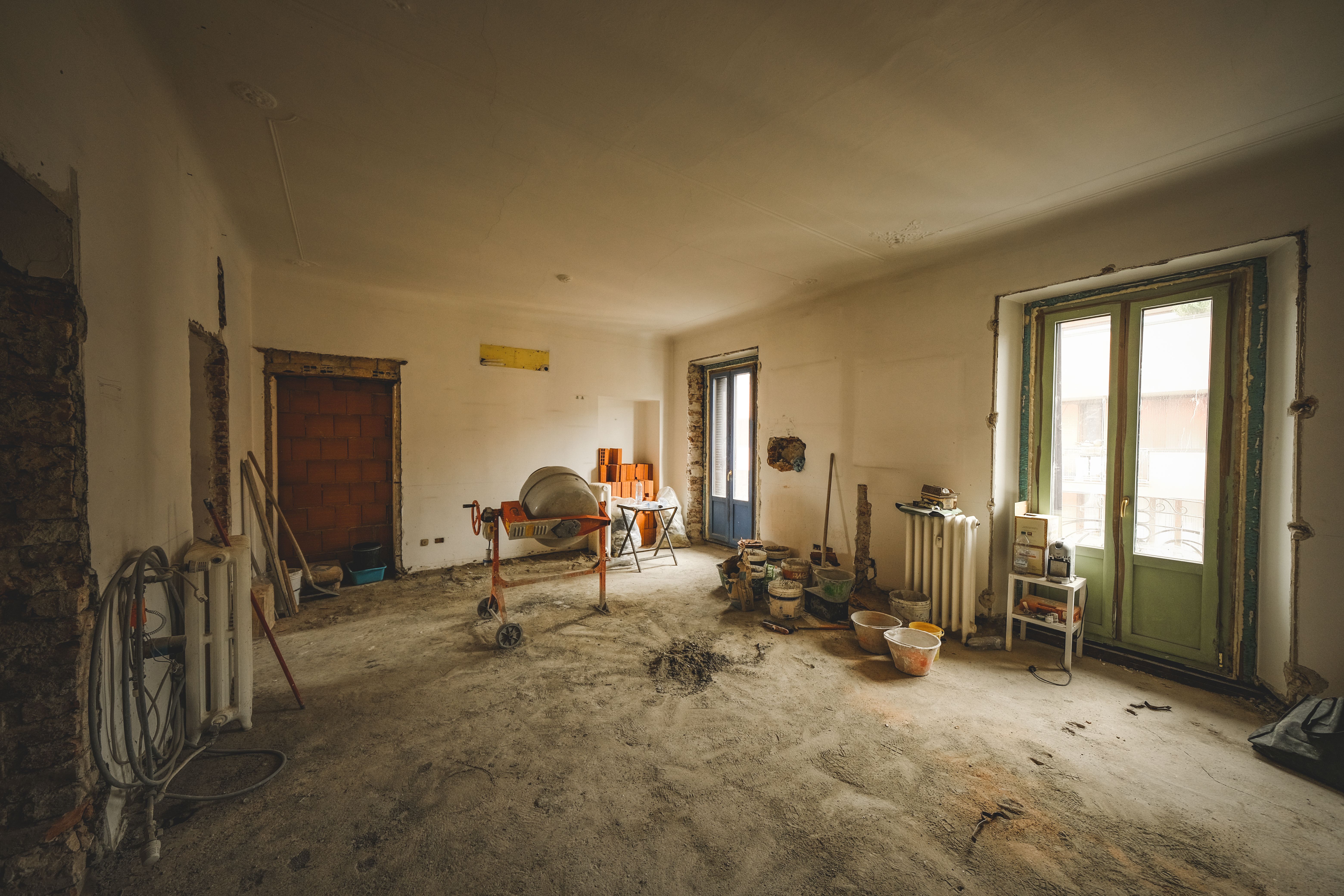 A large room mid-renovation with stripped plaster walls, exposed brick, a cement mixer, and paint buckets on the floor.