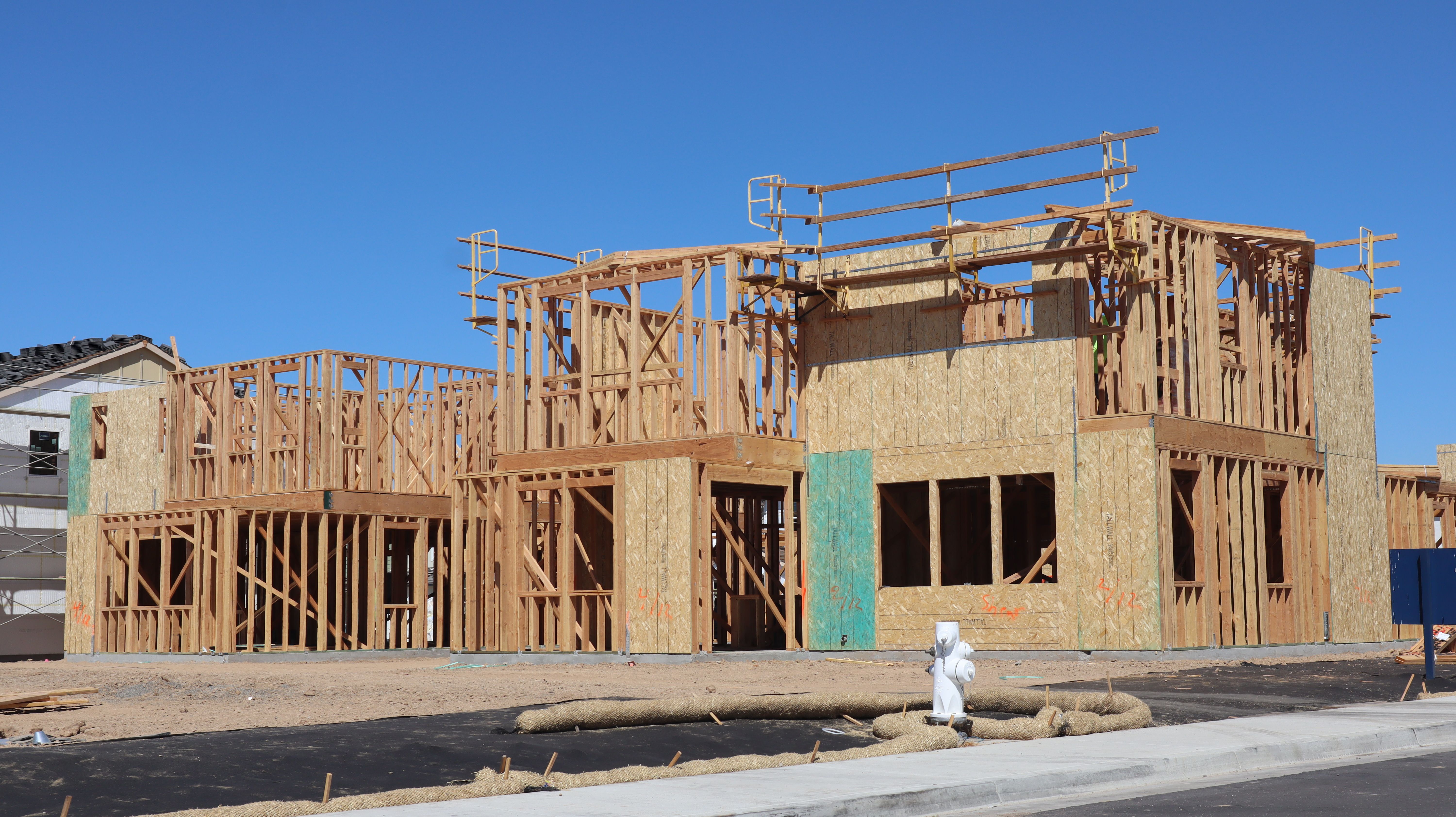 A two-story wood-framed house under construction with exposed studs, OSB sheathing, and green wall panels against a clear sky.