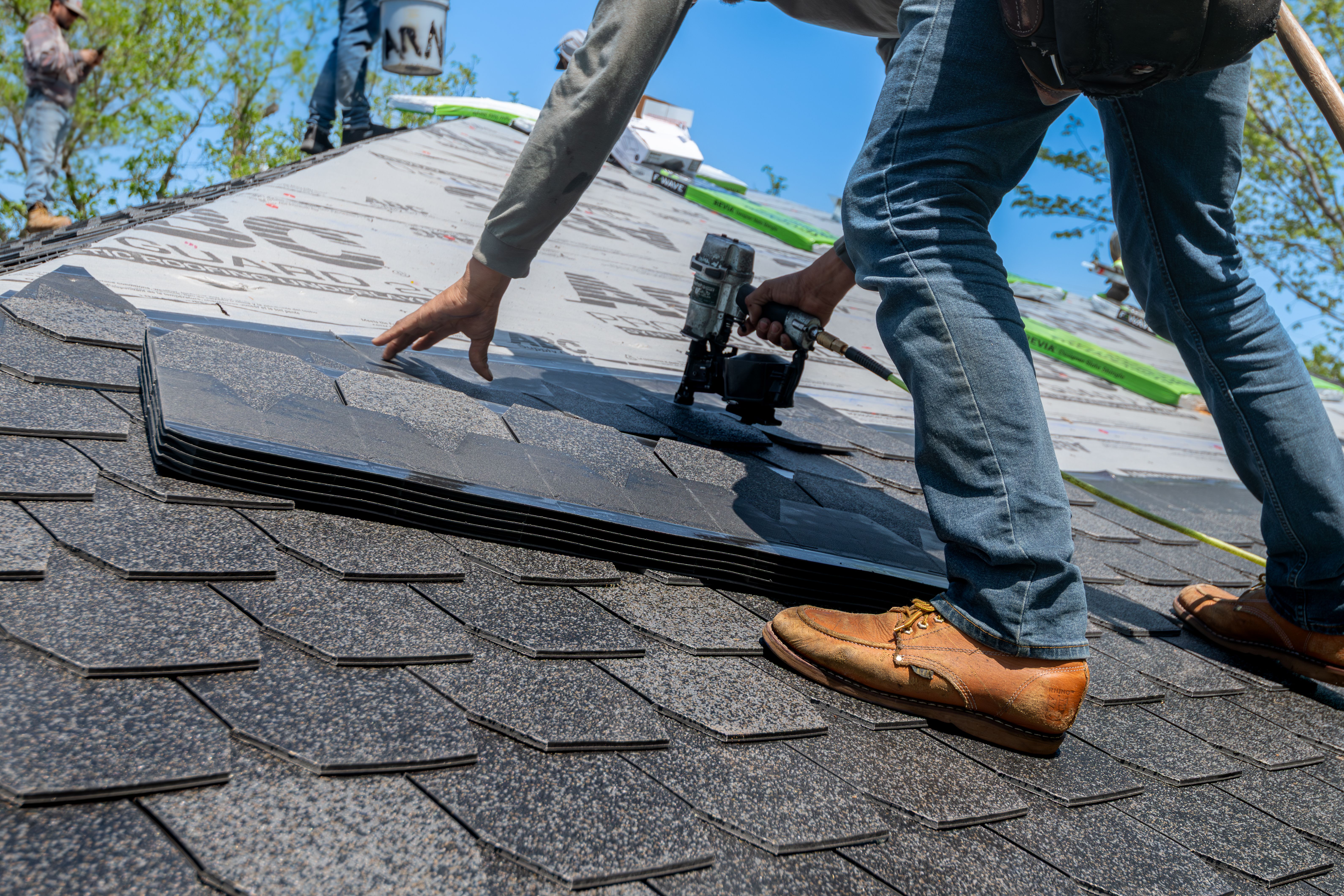 A roofer in work boots uses a pneumatic nail gun to install dark asphalt shingles on a residential roof.