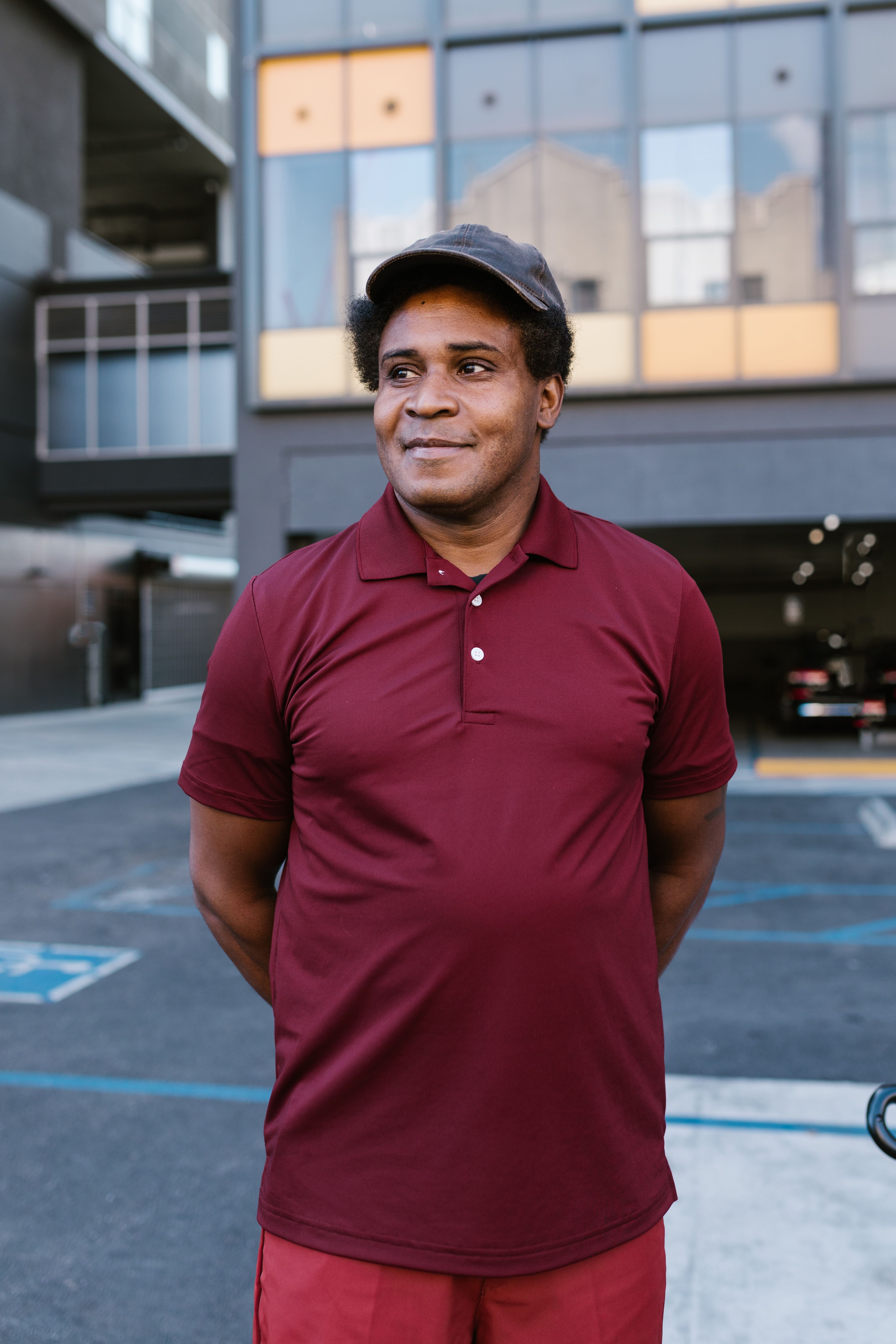 Portrait of a smiling middle-aged man wearing a dark cap and a burgundy polo shirt, standing in a parking lot with a modern building behind him.