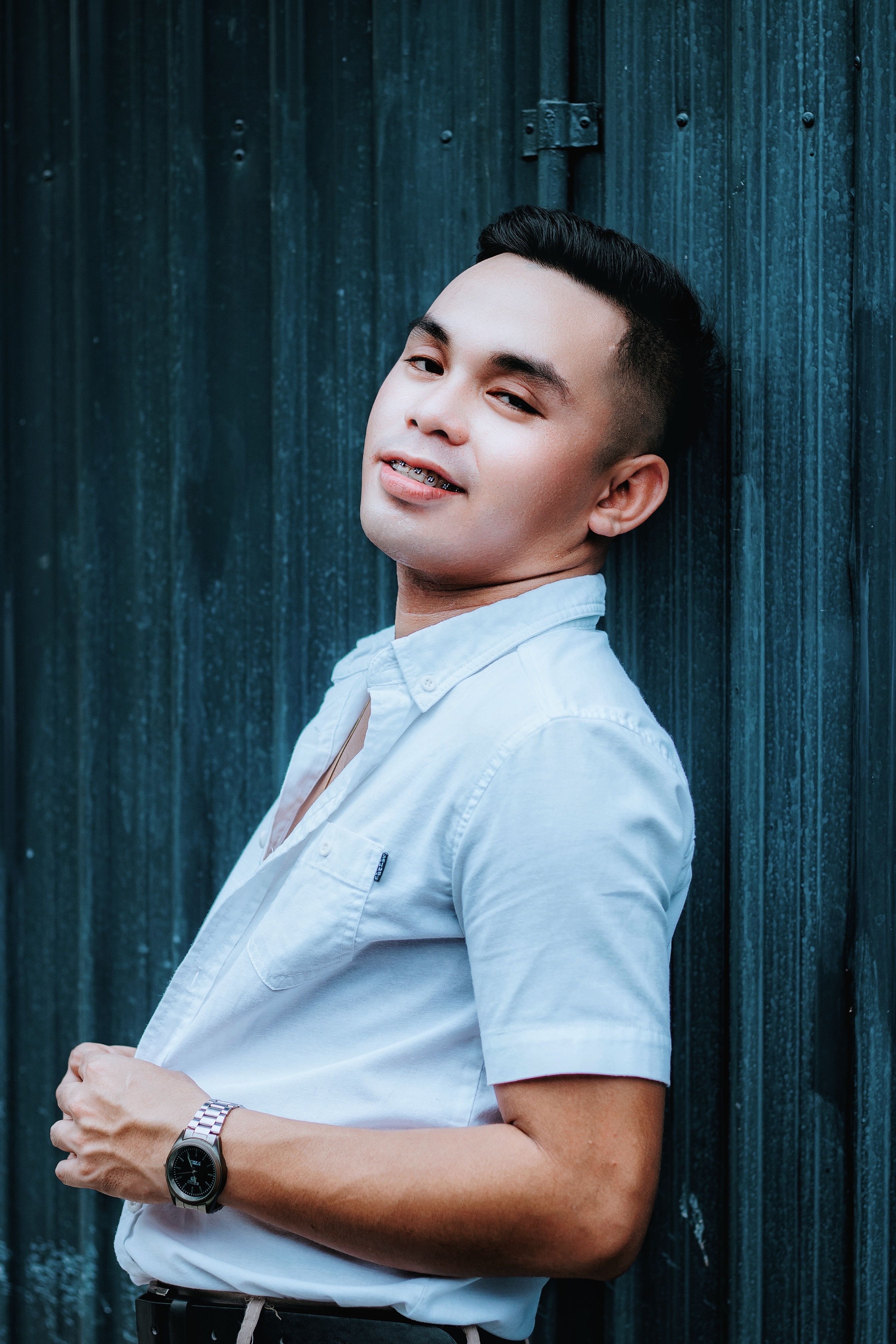 Portrait of a confident young man in a light blue button-down shirt leaning against a dark textured wall.