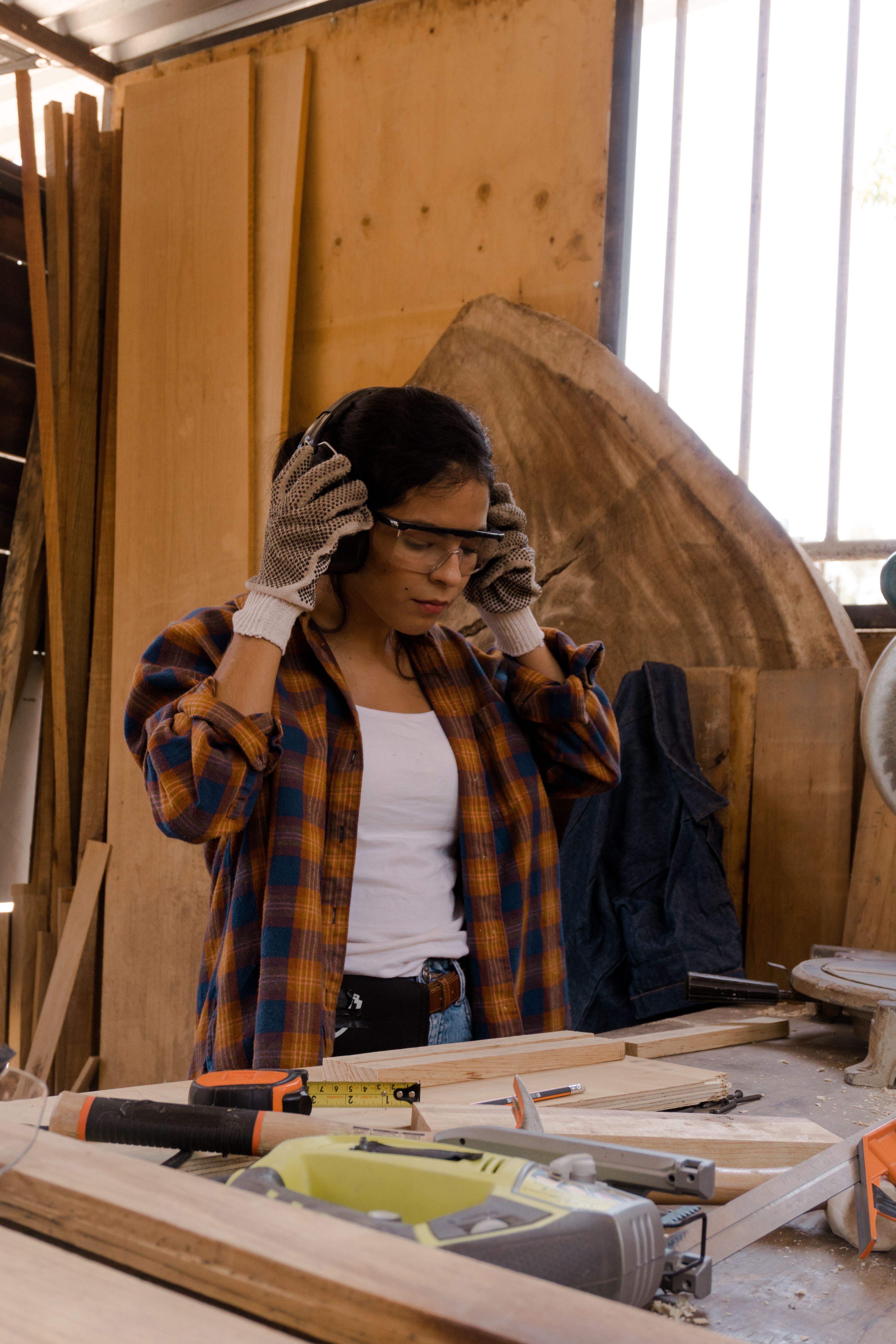Portrait of a young woman in a plaid flannel shirt putting on safety glasses inside a carpentry workshop, with lumber and tools around her.
