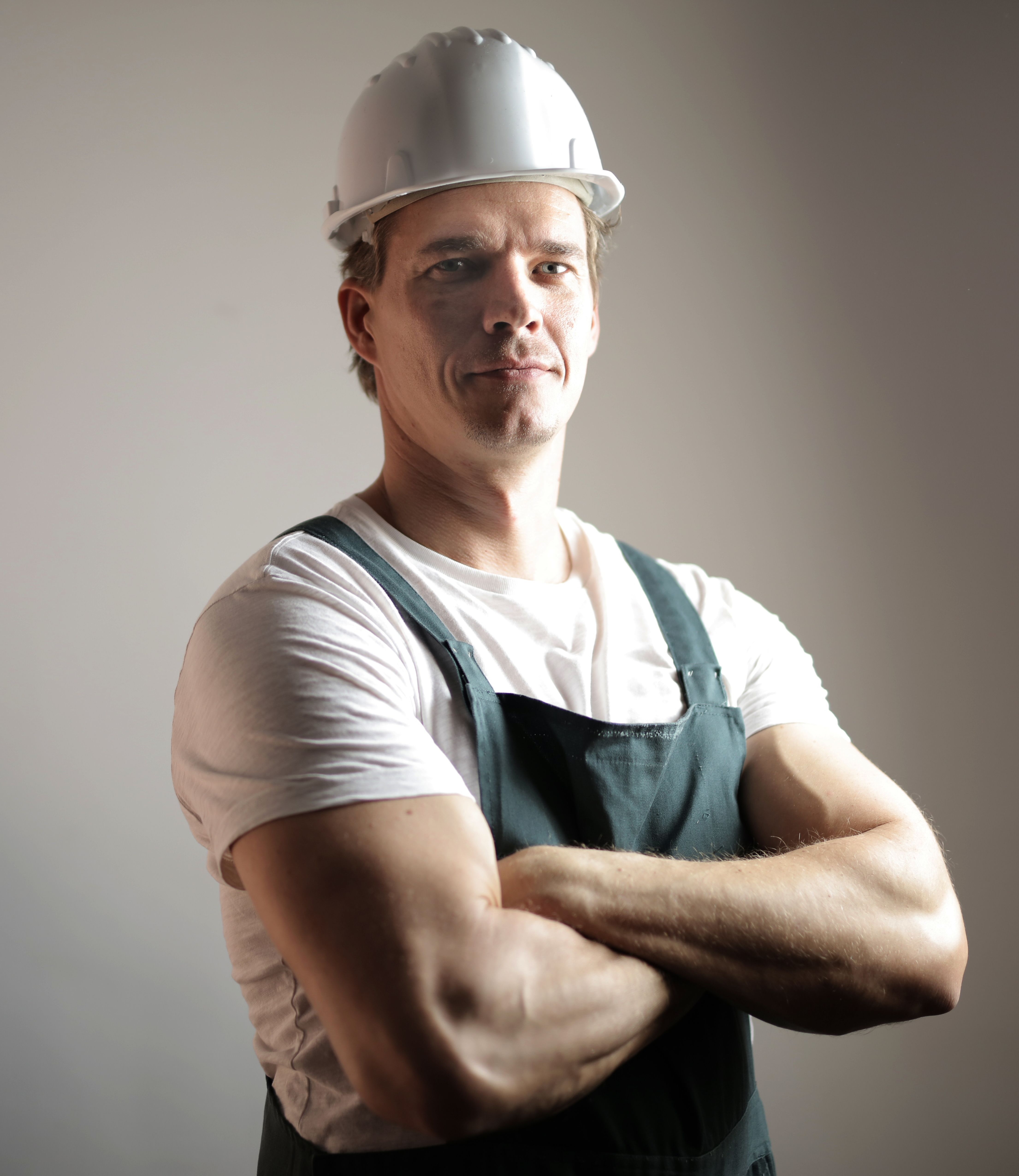Portrait of a confident middle-aged man in a white hard hat, white t-shirt, and dark green work overalls, standing with his arms crossed.