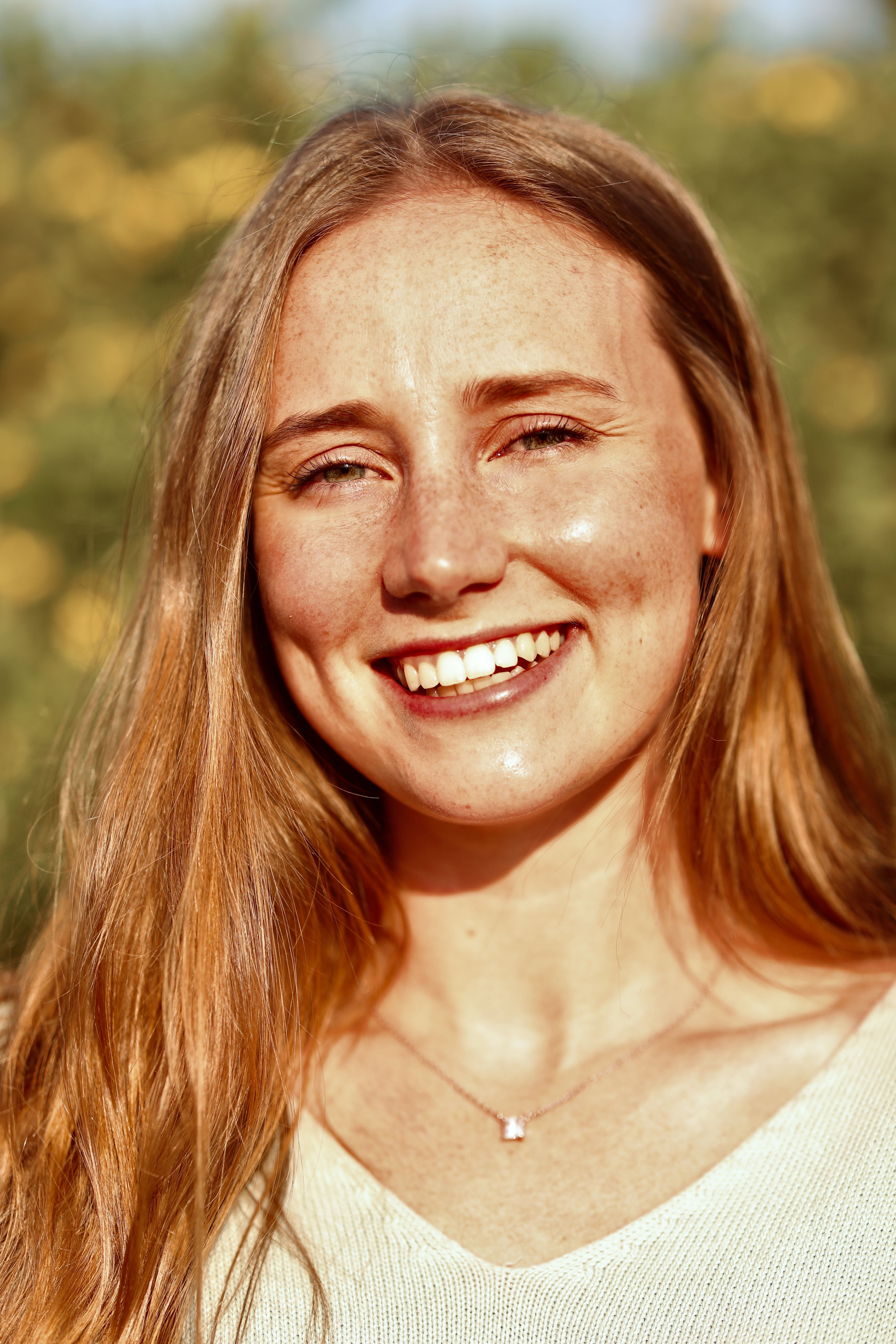 Portrait of a smiling young woman with long hair in a sunlit outdoor setting, with yellow flowers blurred in the background.