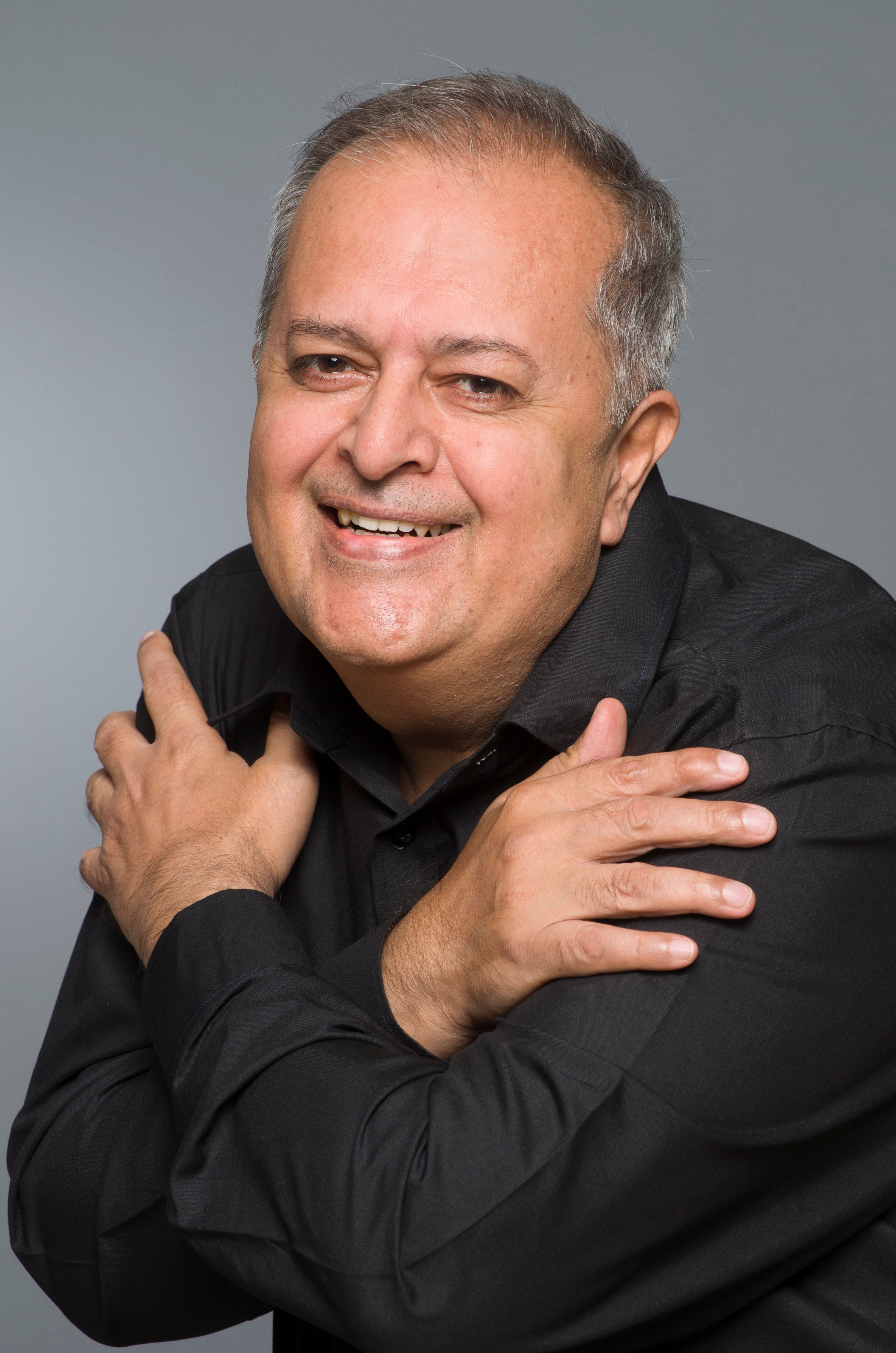 Portrait of a smiling senior man in a black shirt with his arms crossed over his chest against a light gray studio background.