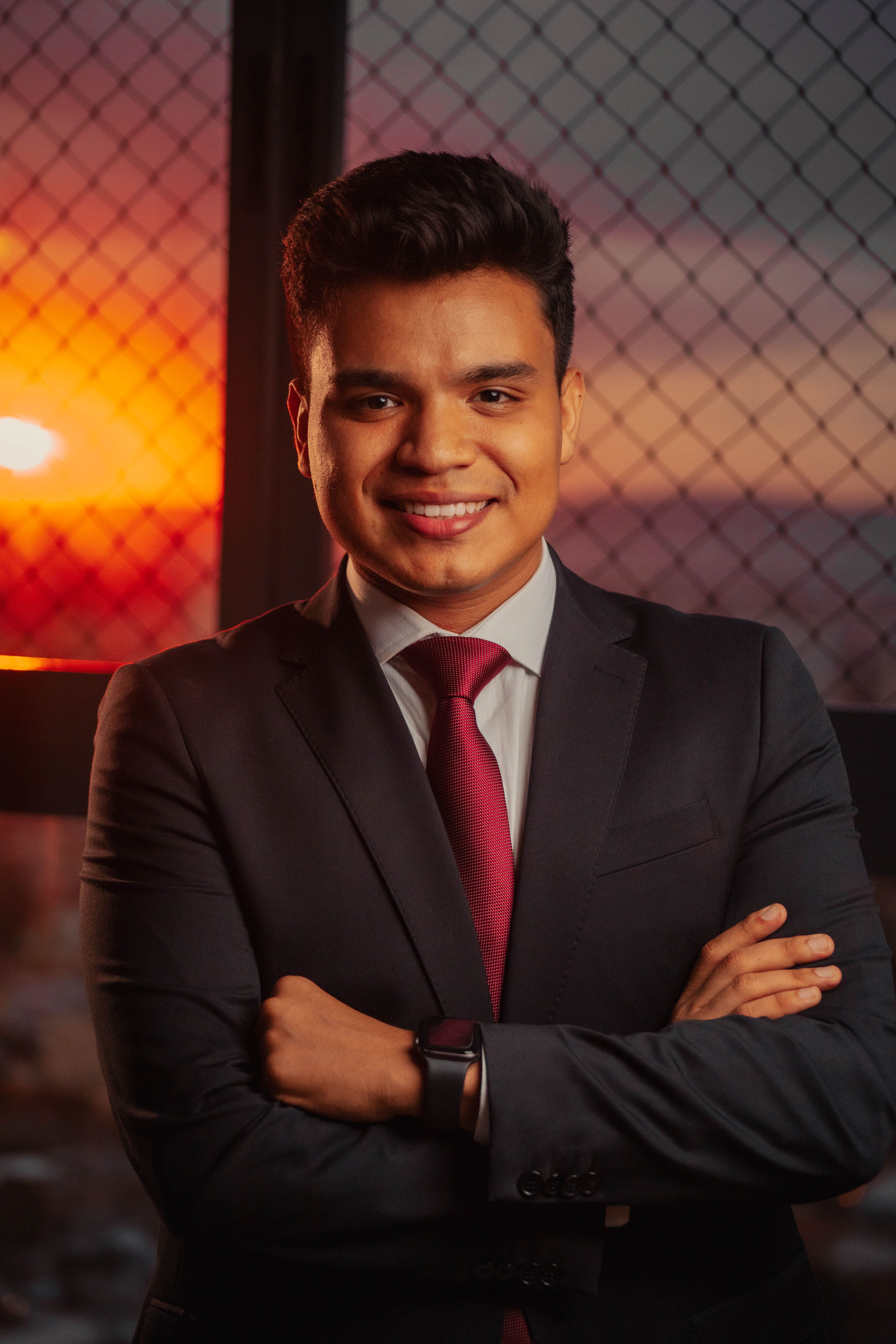 Portrait of a smiling man in a dark suit and red tie with arms crossed, photographed at sunset on a high-rise balcony with a city behind him.