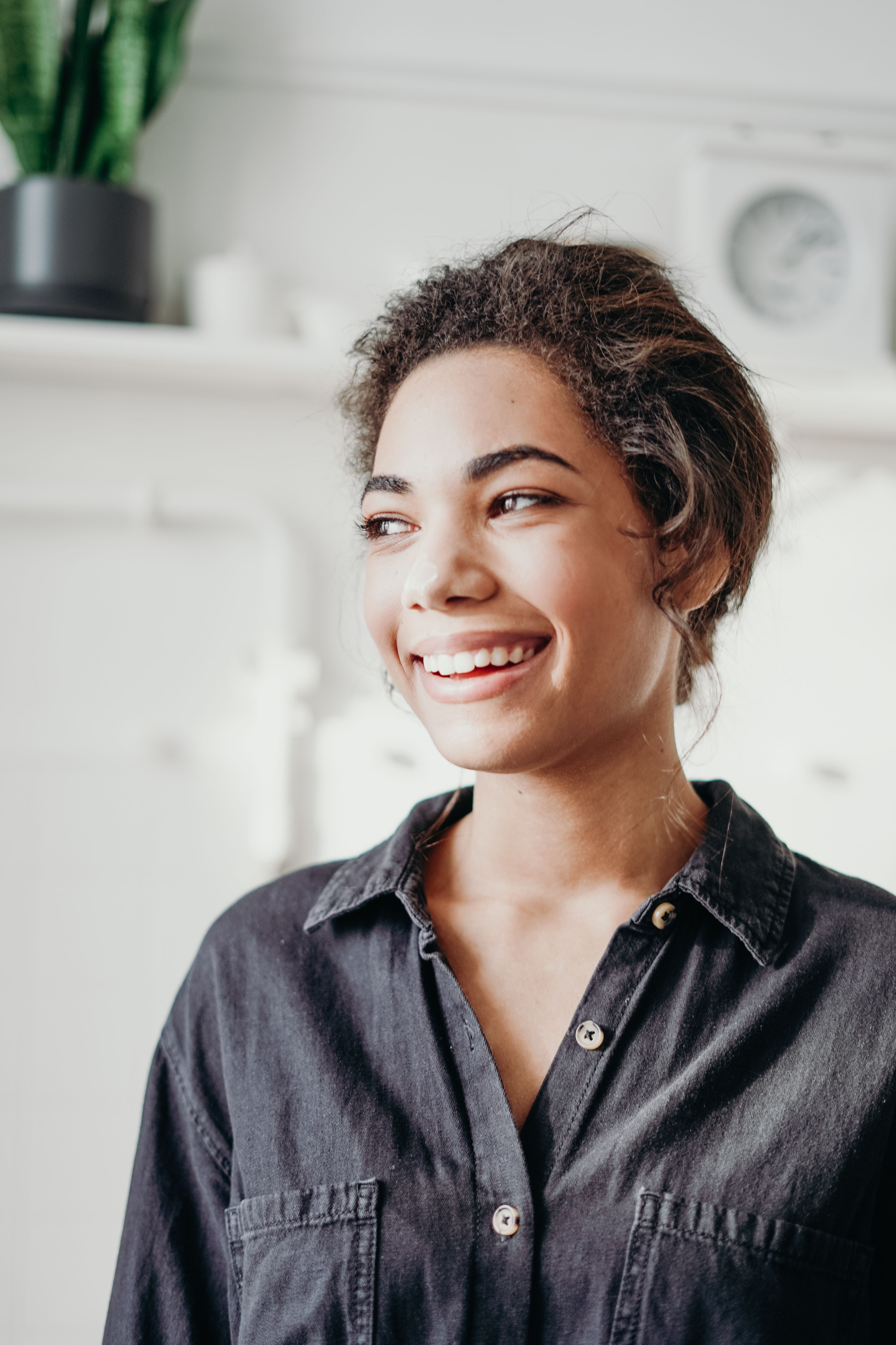 Portrait of a smiling young woman with curly hair in a dark button-down shirt, laughing in a bright indoor home setting.