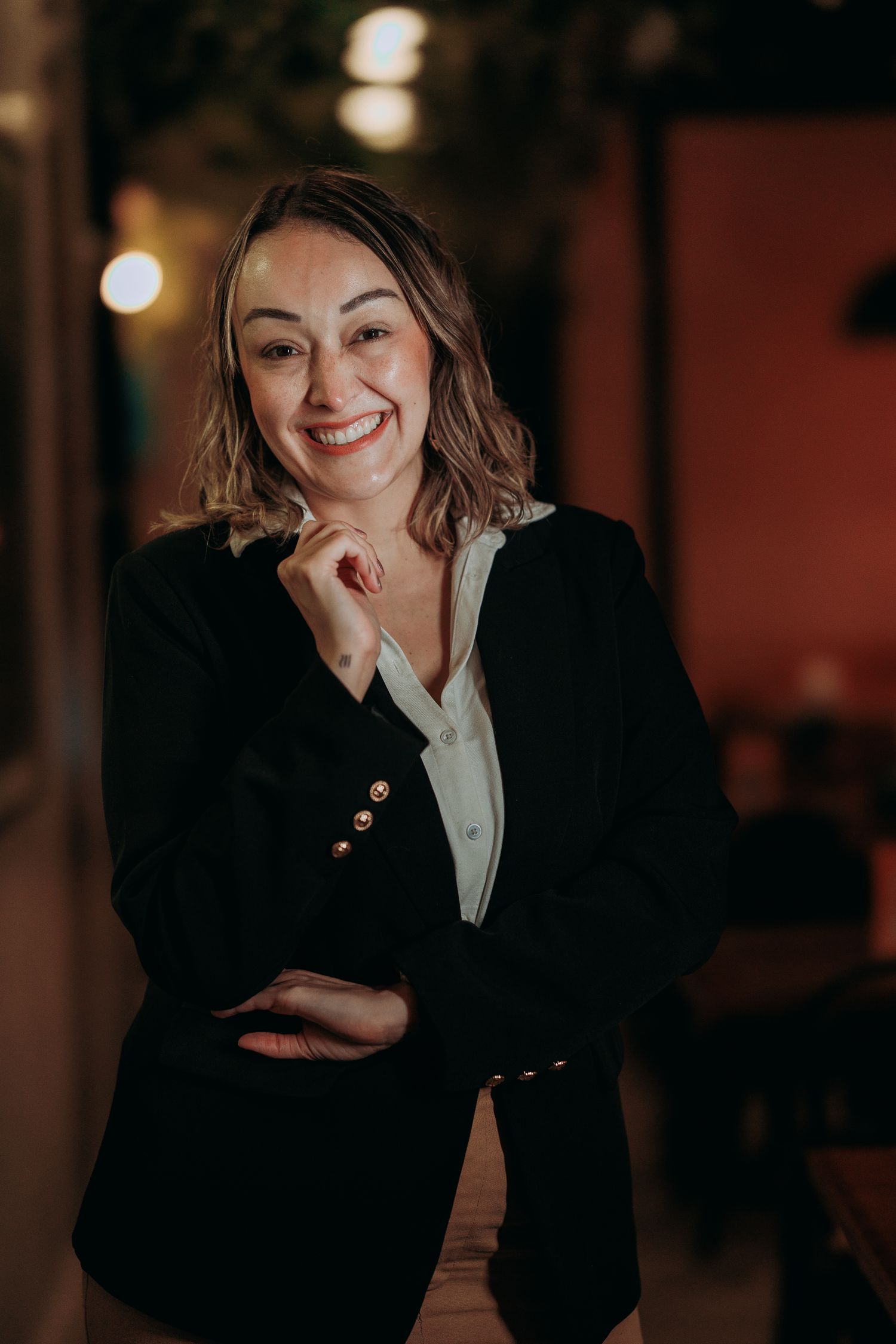 A professional woman with shoulder-length hair smiling in a black blazer and white blouse, arms crossed, in a warmly lit indoor setting.