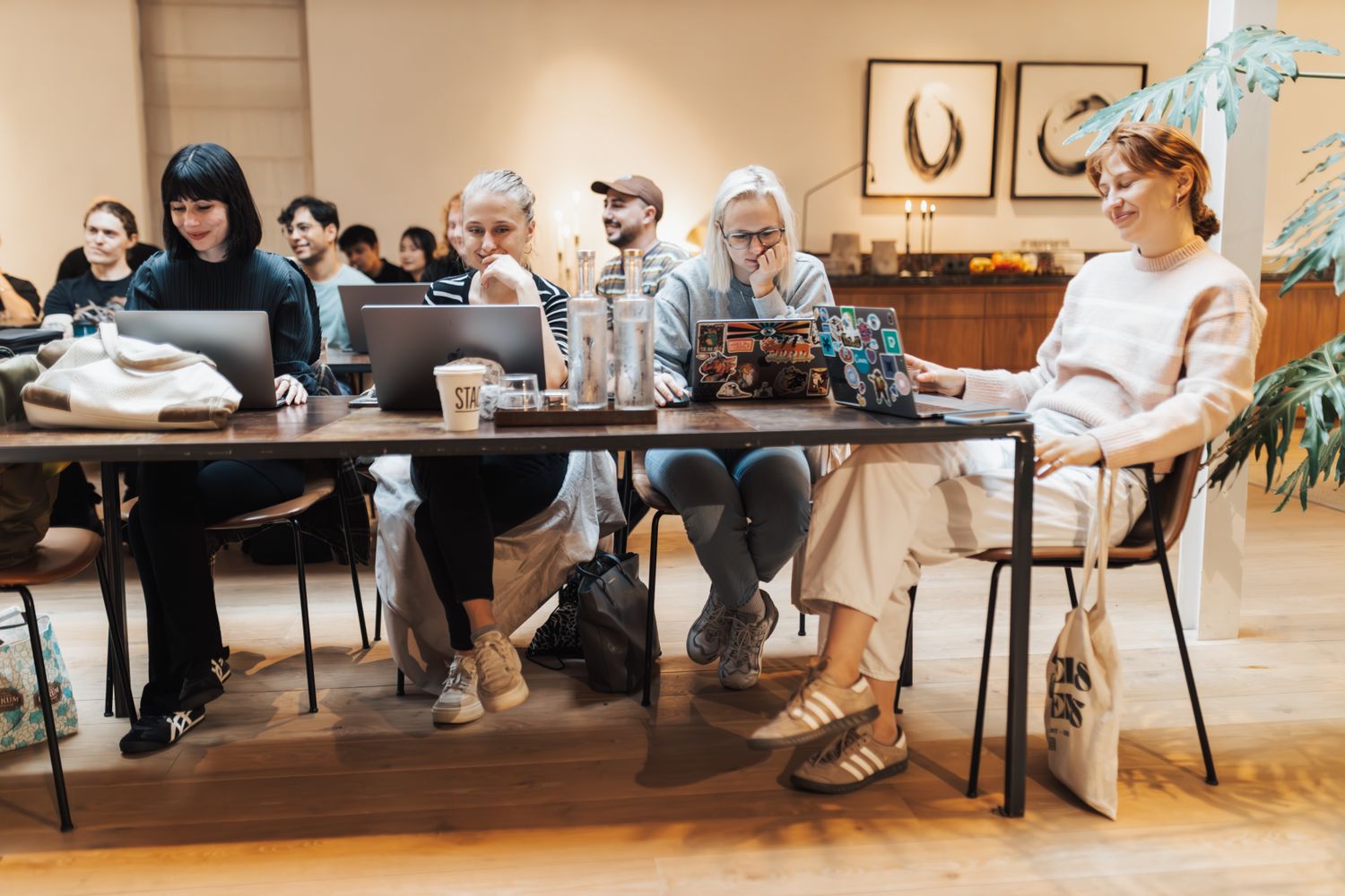A diverse group of adults sitting at a long wooden table working on laptops together in a warm modern space, smiling and engaged.