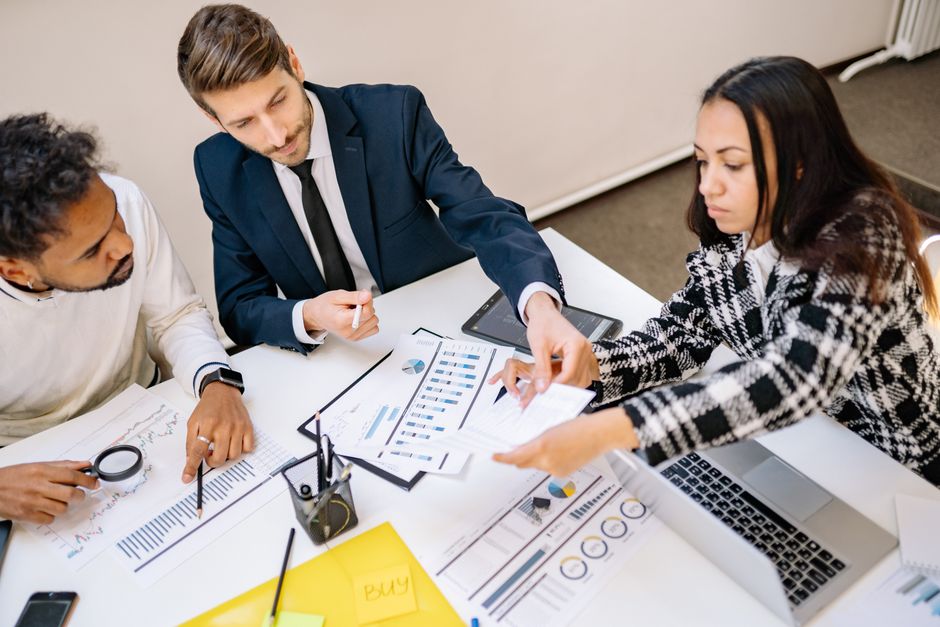 Three business professionals in an office leaning over a table covered with printed charts, a laptop, a magnifying glass, and a yellow notepad.