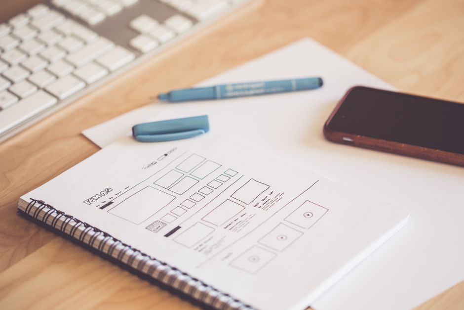 A notebook showing a hand-drawn wireframe of an app layout, with a pen, smartphone, and keyboard on a wooden desk.
