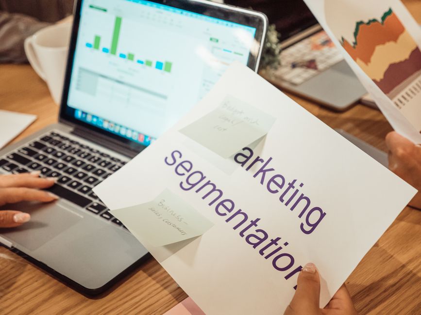 Hands holding a printed marketing segmentation card in front of a laptop showing a bar chart, with a coffee cup and a map printout on the desk.