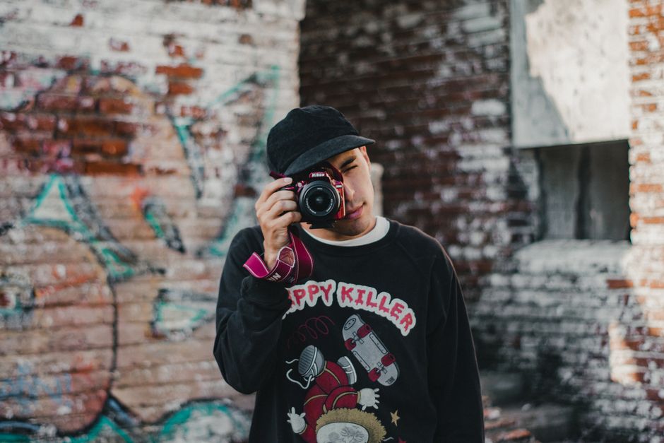 A young man with a black cap holding a DSLR camera up to his face, standing in front of a graffiti-covered brick wall.