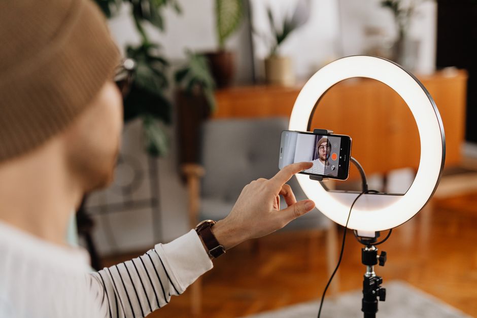 A person filming a video with a smartphone mounted on a ring light and tripod, with the phone screen showing a record interface.