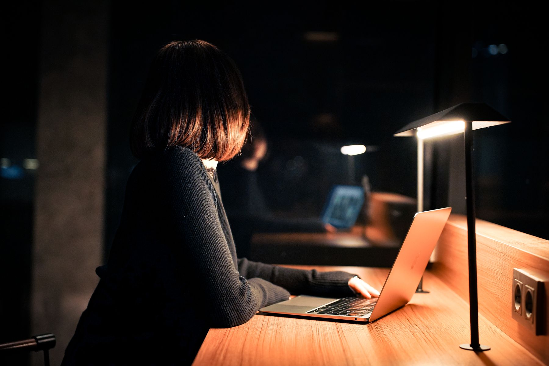 A young woman in a dark blue knit sweater working at a slim silver laptop in a dark, moody nighttime study setting, illuminated by a warm table lamp on a long wooden counter.