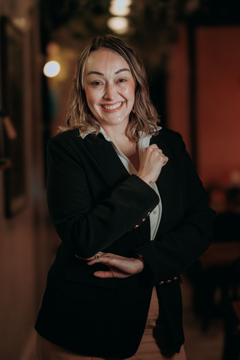 A woman with shoulder-length light-brown hair wearing a black blazer and white blouse, bright smile, professional headshot with warm bokeh background.