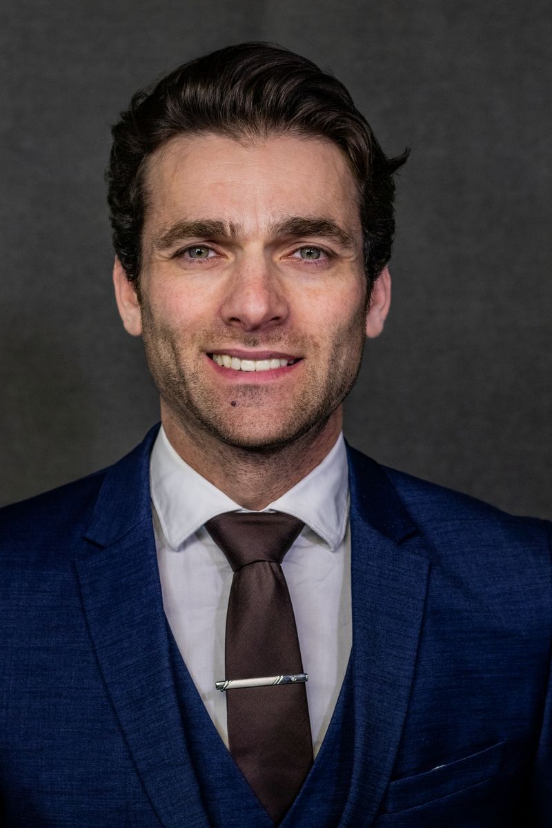 A smiling man with short brown hair and slight stubble wearing a navy blue suit and white shirt, professional headshot against a dark gray background.