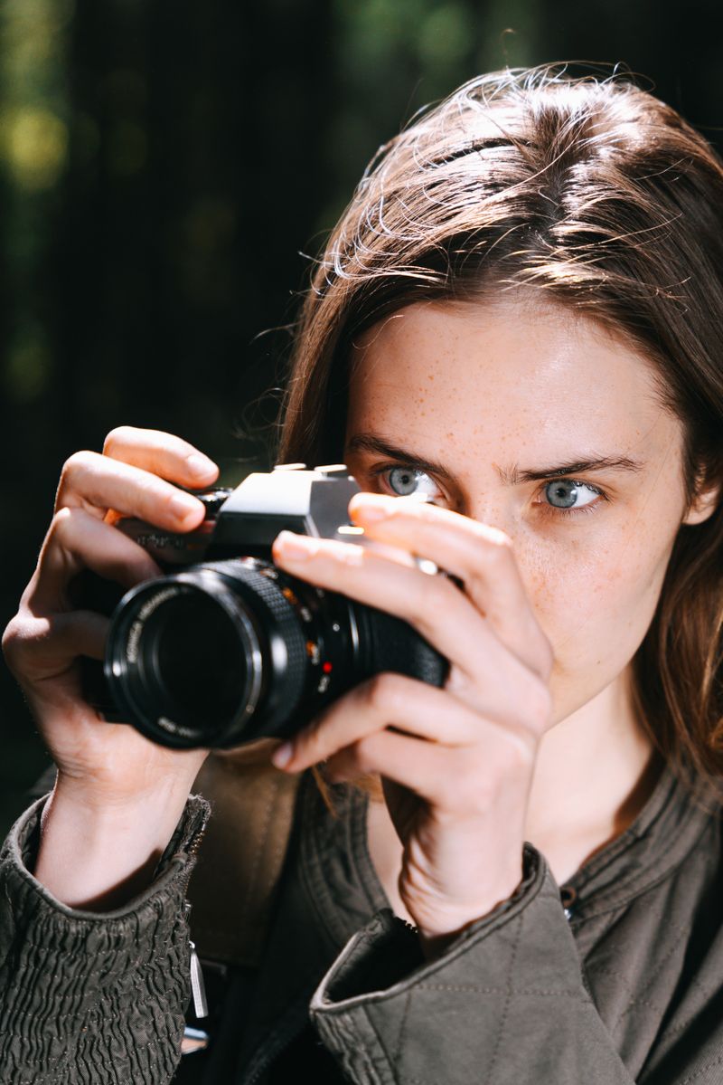 A woman with long light-brown hair holding a camera up to her eye, focused expression, professional portrait with a softly blurred outdoor background.