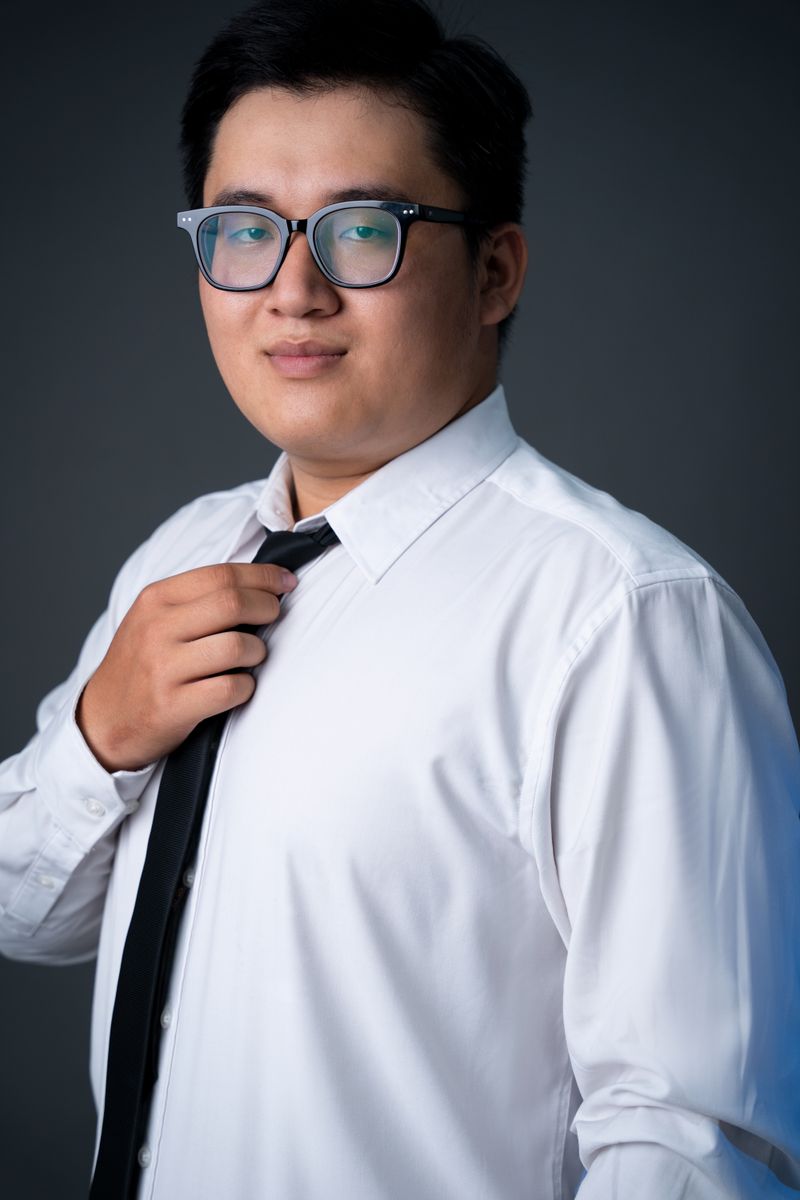 A man with short black hair and glasses wearing a white dress shirt and adjusting a black tie, professional headshot against a dark gray background.