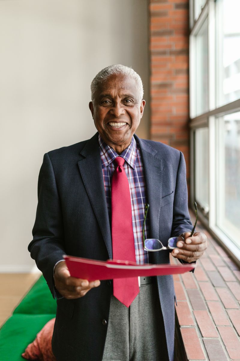 An older man with short gray hair wearing a navy blazer and plaid shirt, warm smile, holding glasses and a folder, professional portrait in front of a brick wall and window.