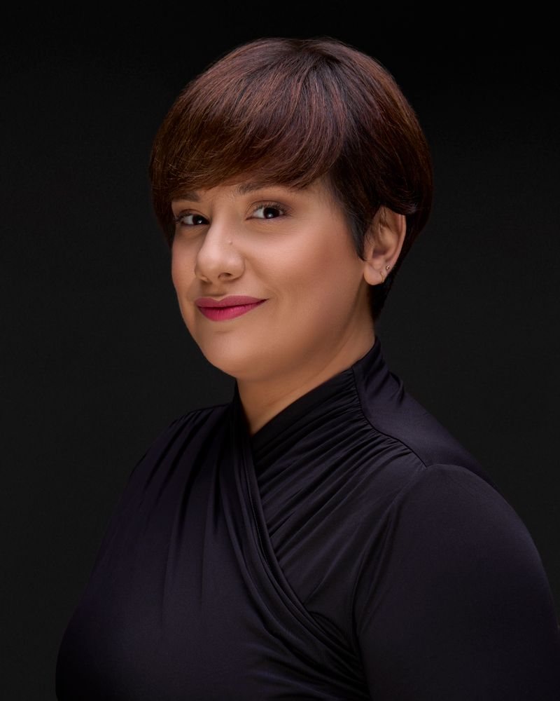 A woman with short reddish-brown hair wearing a black draped top, soft smile, professional headshot against a dark background.