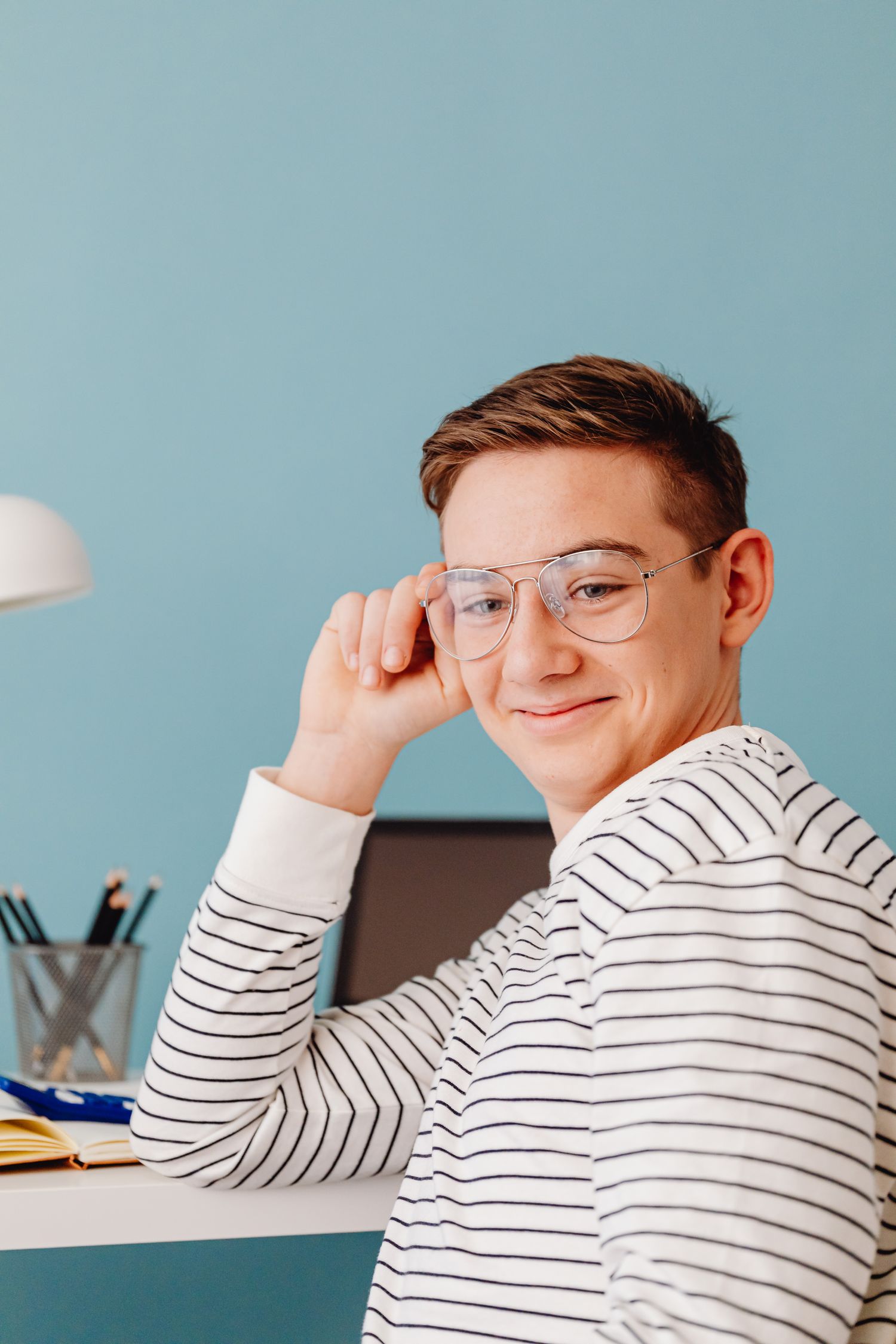 A young man with glasses and a striped shirt sitting at a desk with pencils and an open notebook, slight smile against a light blue background.