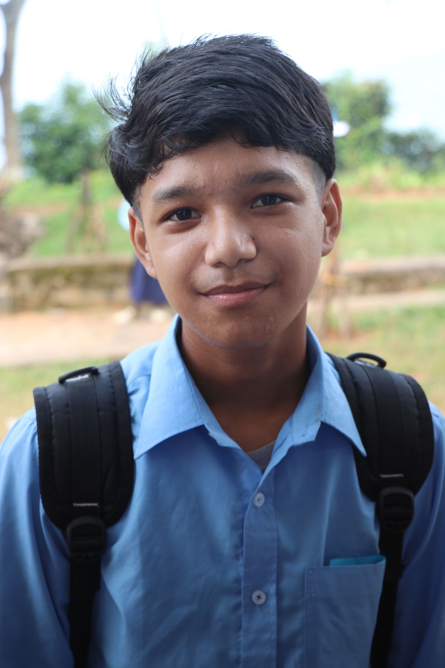 A young man wearing a blue button-down shirt and a backpack, slight smile outdoors with a green background.