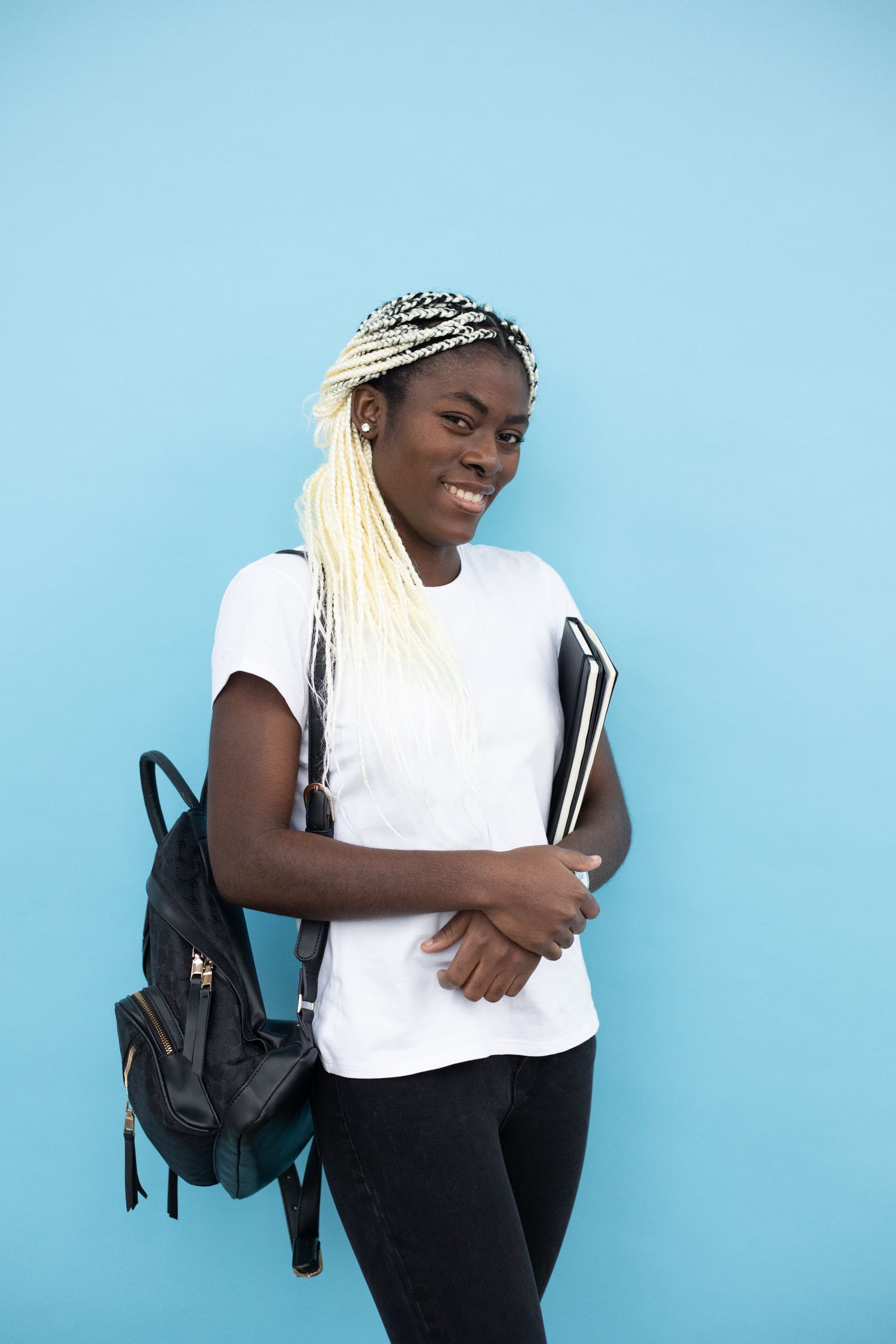 A young woman with long blonde braids wearing a white t-shirt and backpack, holding notebooks, smiling against a bright blue background.