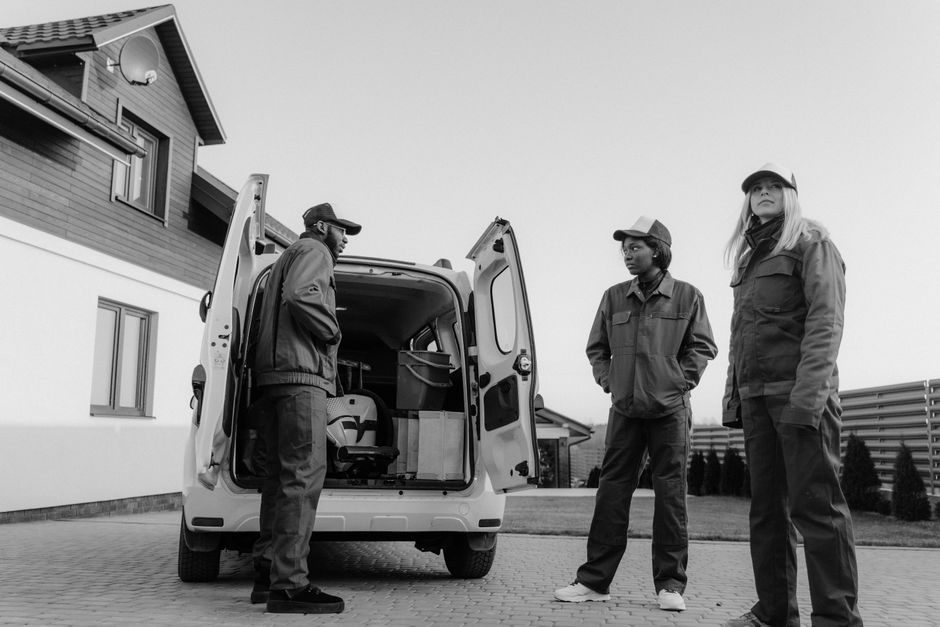 Three workers in matching grey coveralls stand together by the open rear doors of a white work van in a residential courtyard, in black and white.