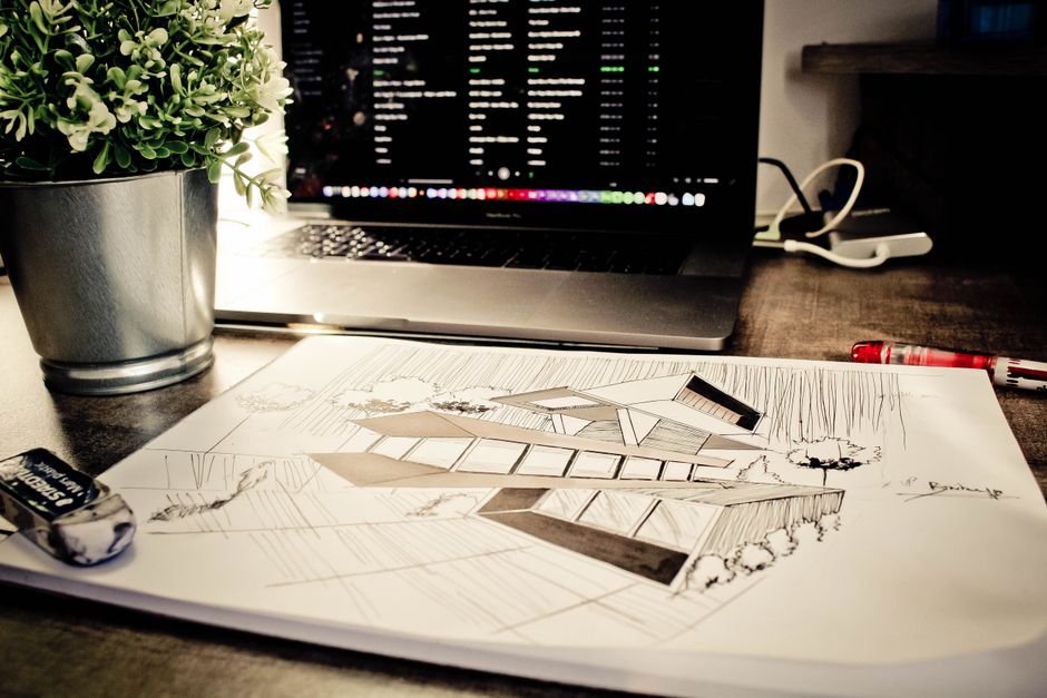 A hand-drawn perspective architectural sketch of a modern outdoor patio lies on a wooden desk beside a small potted plant, an open laptop, and drawing pens.