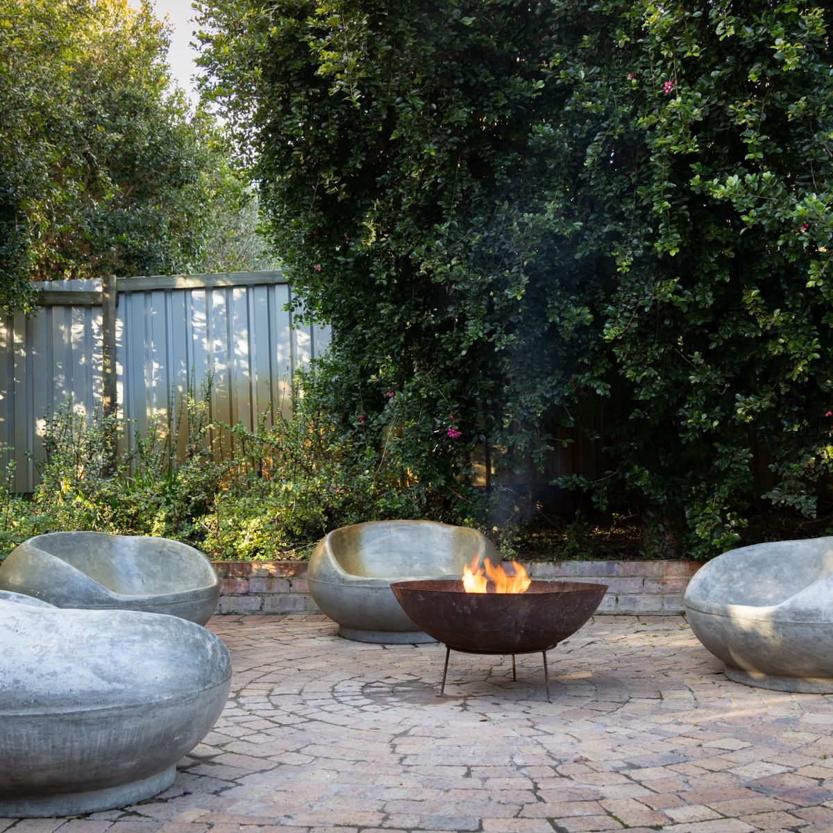 A circular stone patio with smooth concrete pod chairs gathered around a small lit fire pit, backed by a tall flowering hedge and a metal fence.