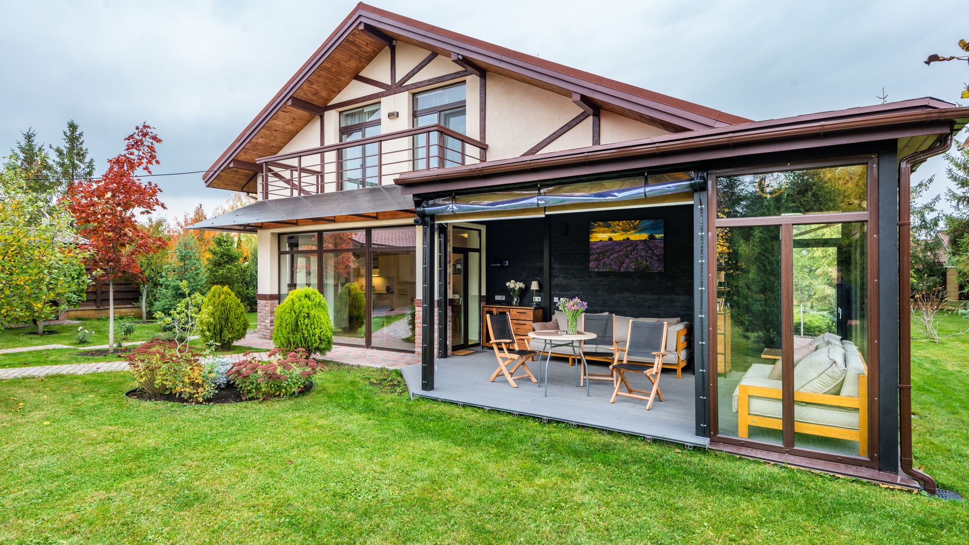 A two-story suburban house with a glass-enclosed modern patio extension, set on a large green lawn with a perennial flower bed and a red Japanese maple.