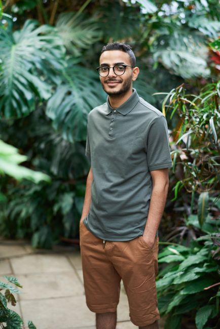 A smiling man in a sage-green polo shirt and glasses stands in a lush tropical garden, hands in pockets, looking warmly at the camera.