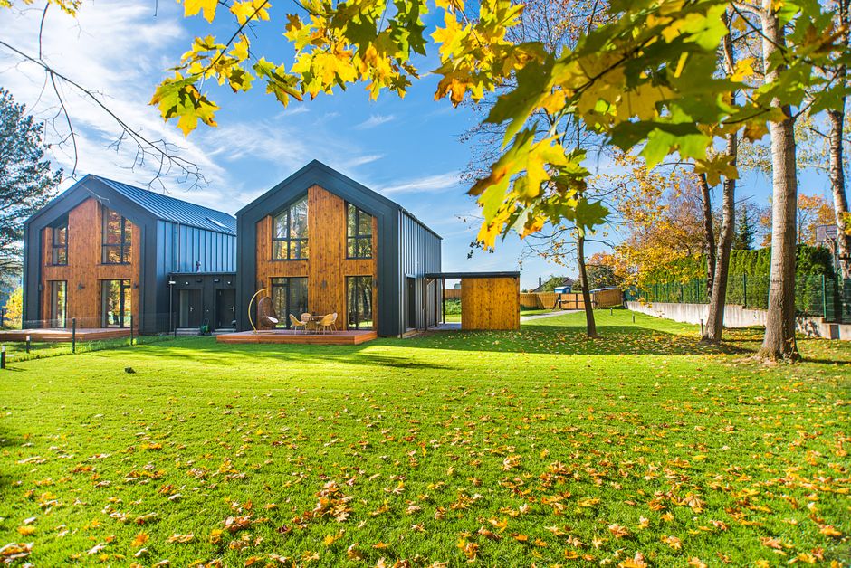 Two modern dark-wood contemporary houses sit on a lush green lawn scattered with yellow autumn leaves, framed by a brilliant yellow maple tree.