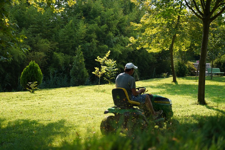 A man in a white cap and grey t-shirt rides a green-and-yellow ride-on lawn mower through a lush green summer lawn, with grass clippings flying in the sun.