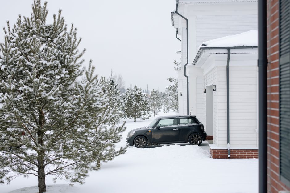 A black Mini Cooper is parked in a snow-blanketed residential driveway beside a white house and a tall snow-laden blue spruce, in deep winter snow.