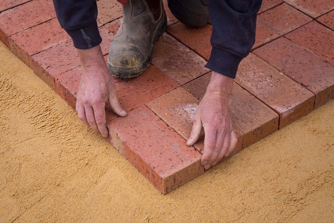 A construction worker's hands carefully place a red brick paver into a partially finished patio pattern bedded on tan sand.