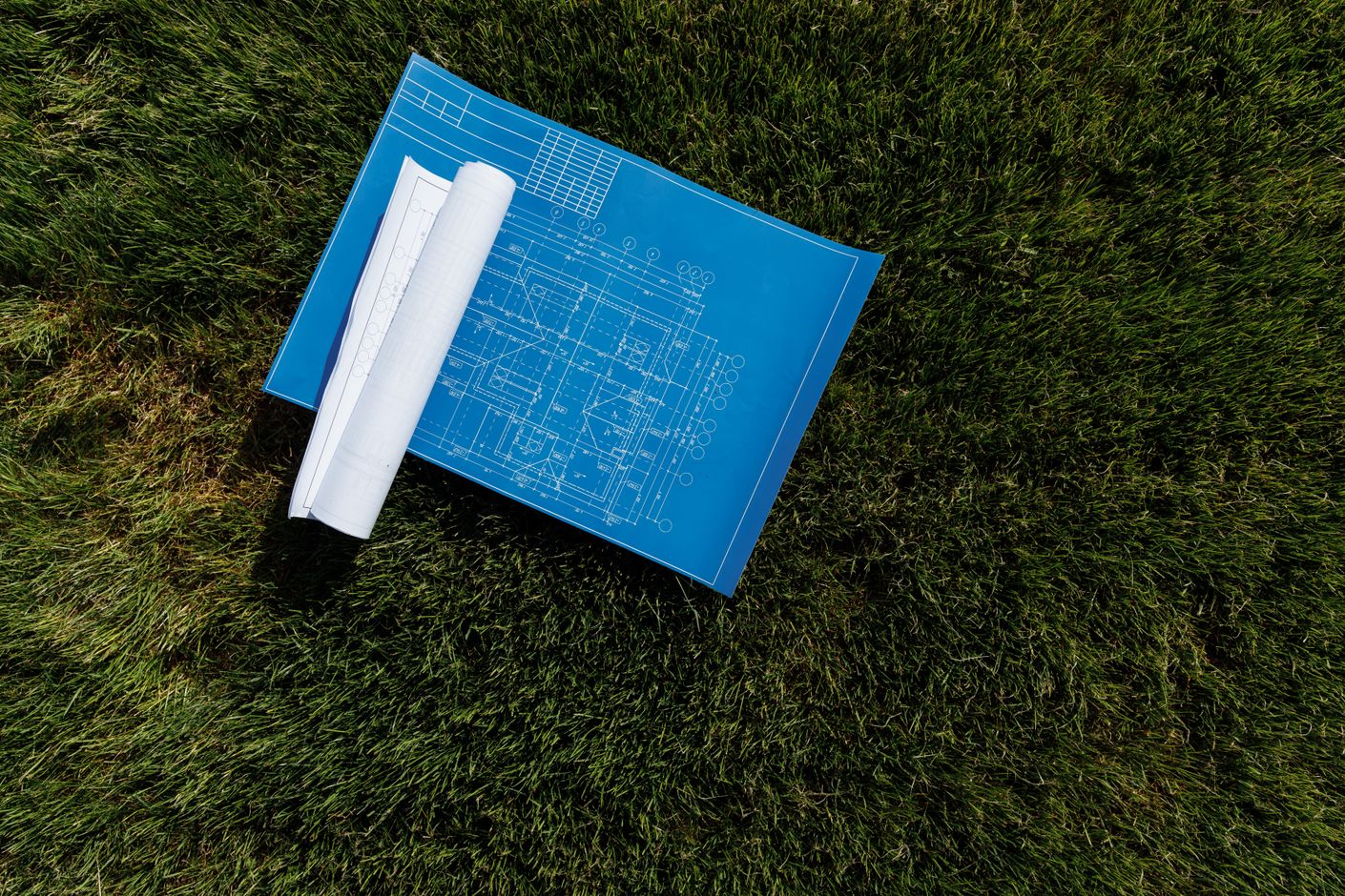 A top-down view of cyan-blue architectural blueprints unrolled on a lush green grass field, with a paper roll of additional plan drawings beside them.
