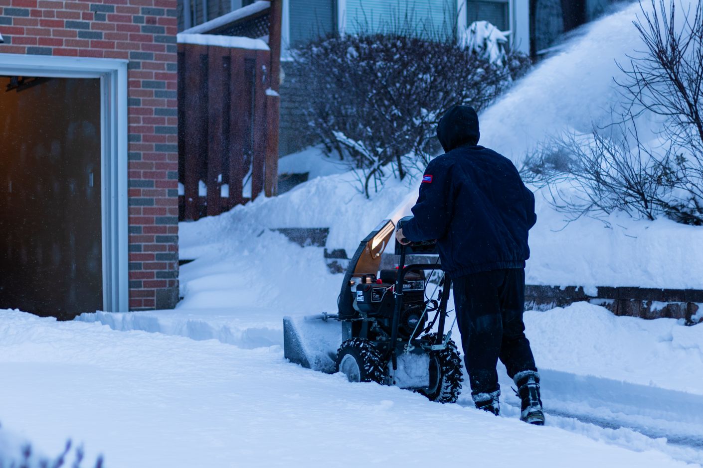 A person in a navy winter jacket uses a snow blower to clear a residential driveway beside a brick house during a heavy snowfall.