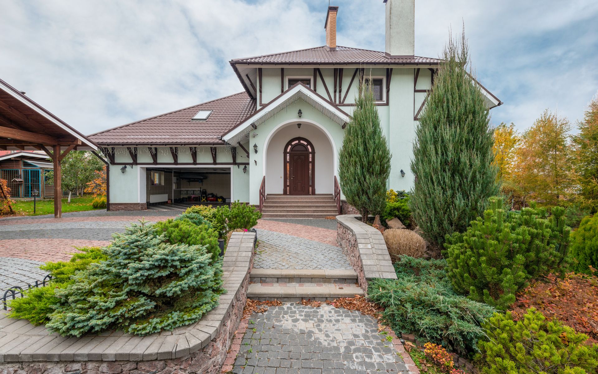 A two-story mint-green house with a brown arched entry, framed by conifers, junipers, and a stone-bordered cobblestone walkway leading to the front door.